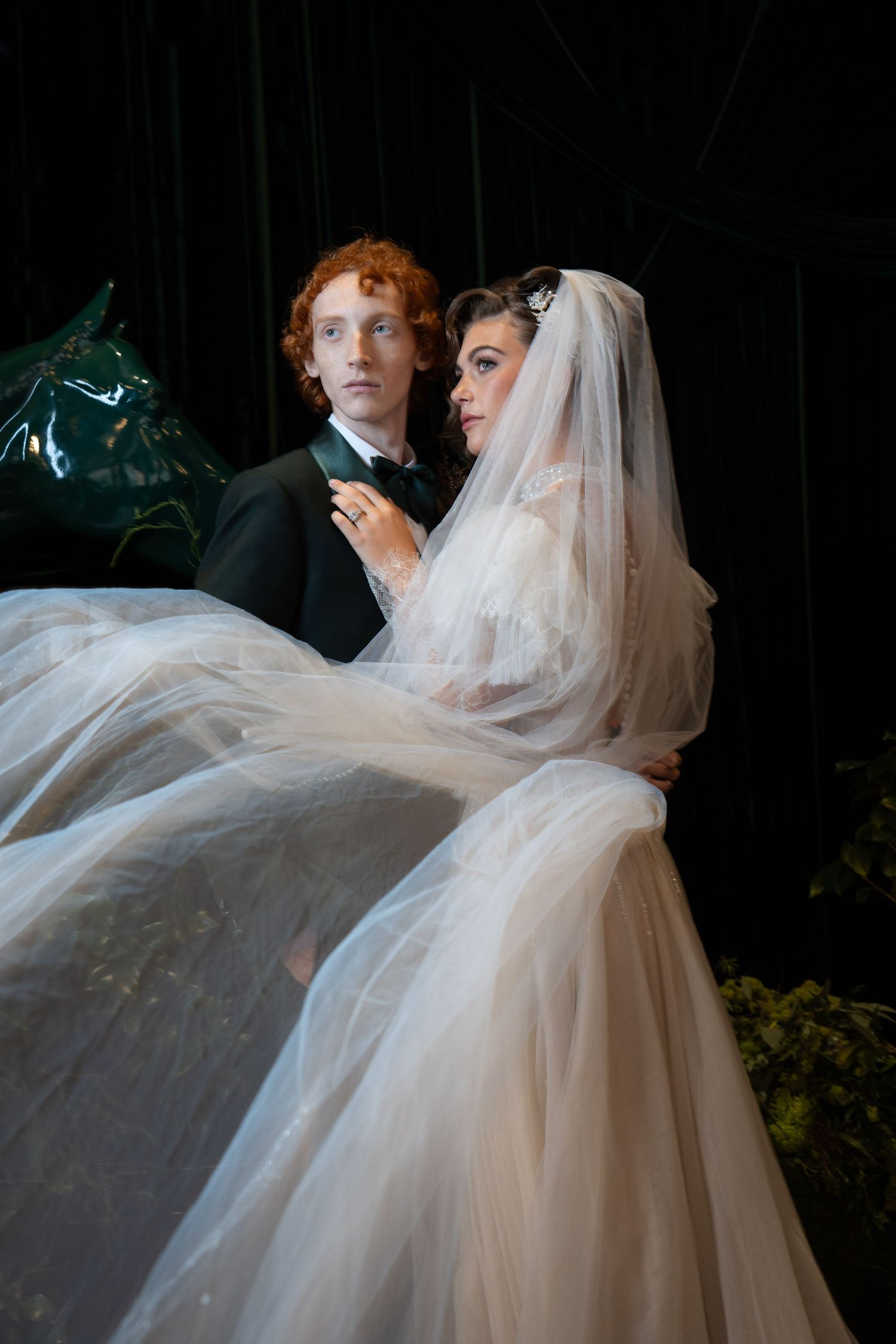 Bride in flowing tulle wedding dress with veil poses with red-haired groom in black tuxedo against dark backdrop.