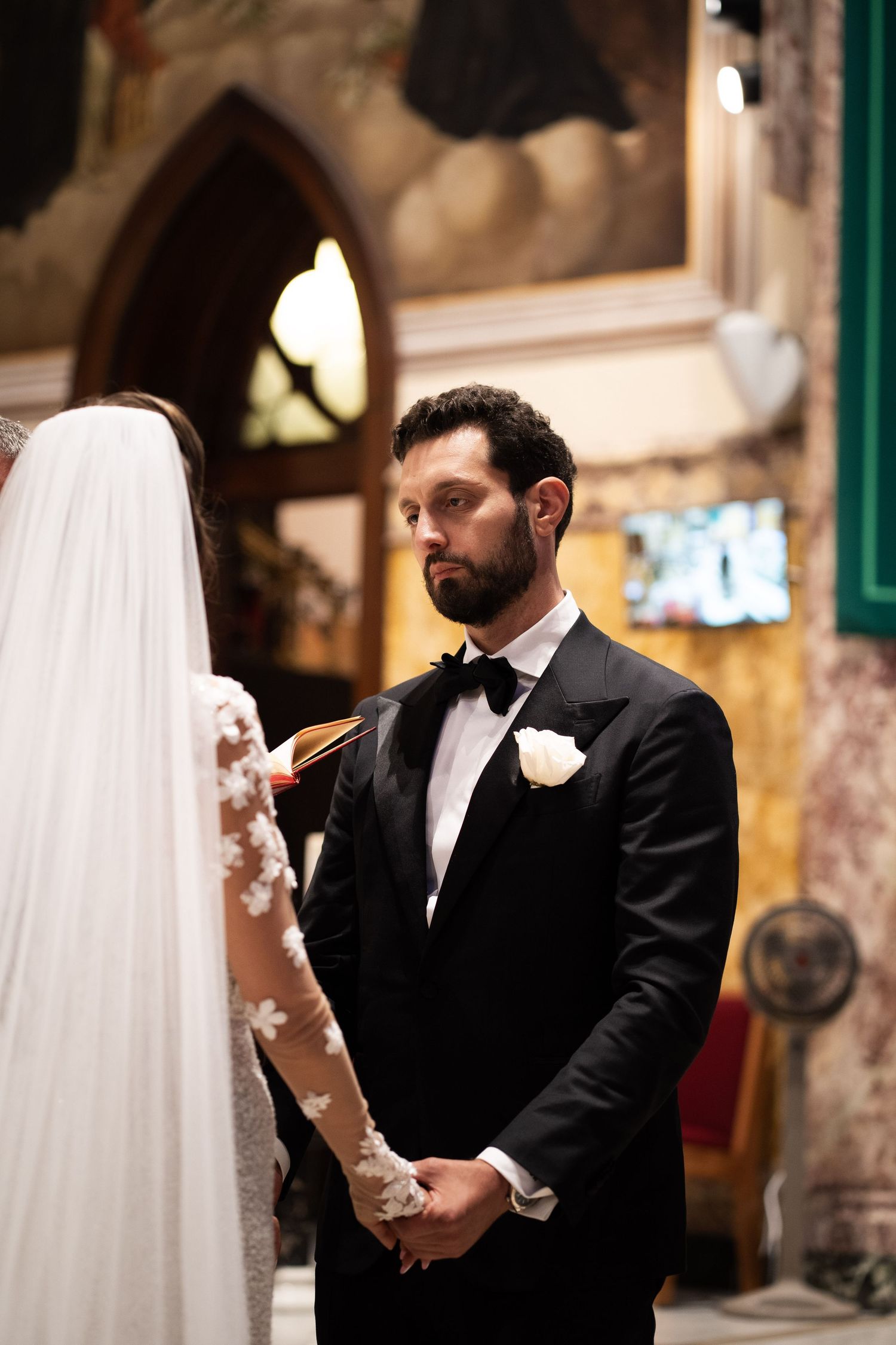 Bearded groom in black tuxedo gazes at bride during emotional wedding ceremony inside historic church.