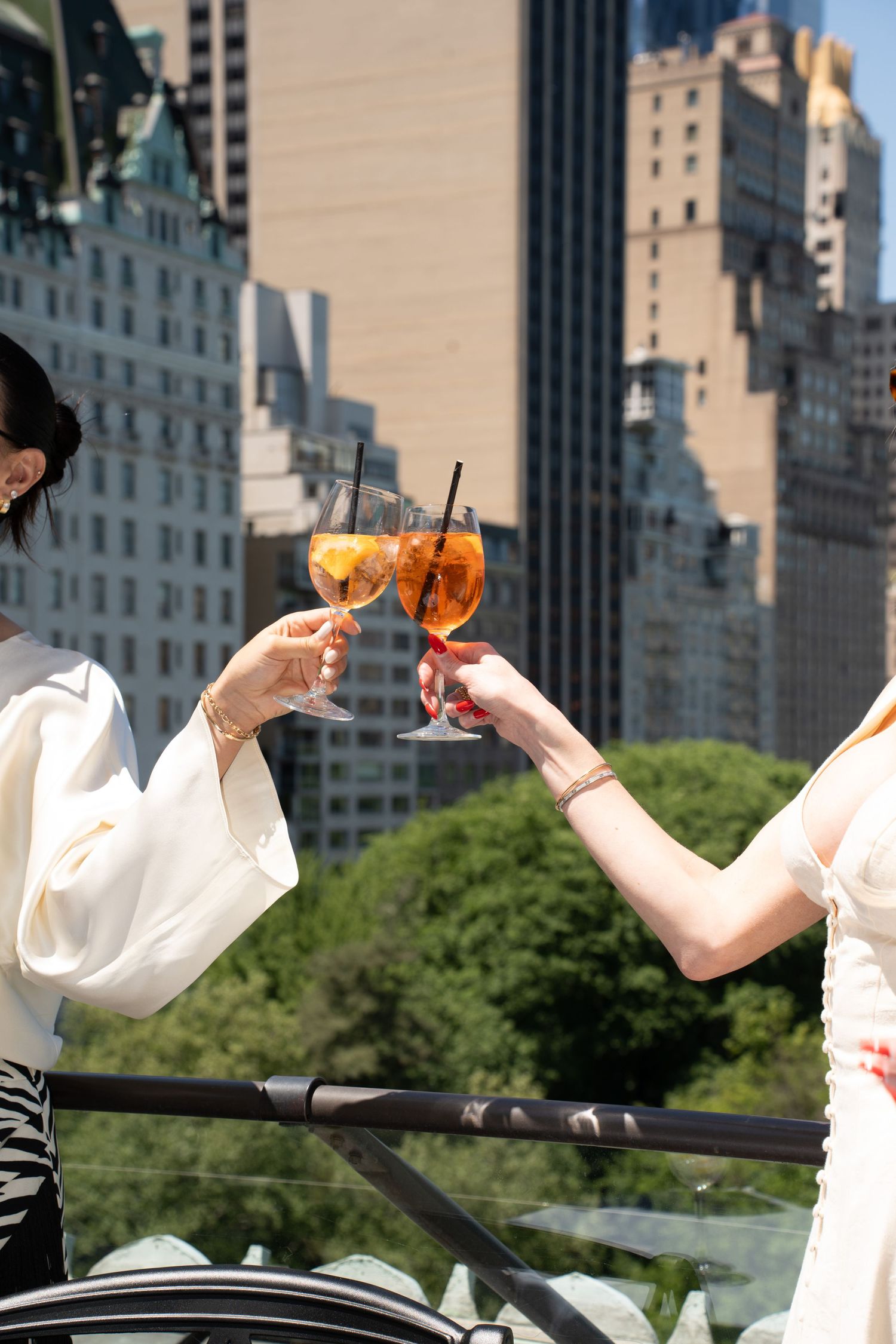 Two women clinking Aperol Spritz glasses on a sunny NYC rooftop with skyscraper backdrop.