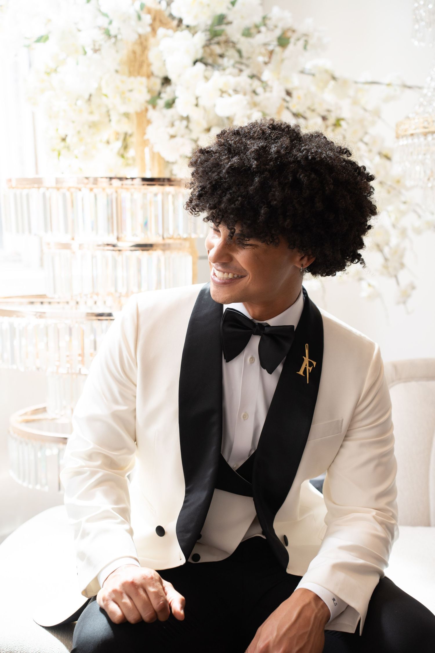 Laughing groom in white tuxedo with black bow tie looks away amid white floral wedding decorations.