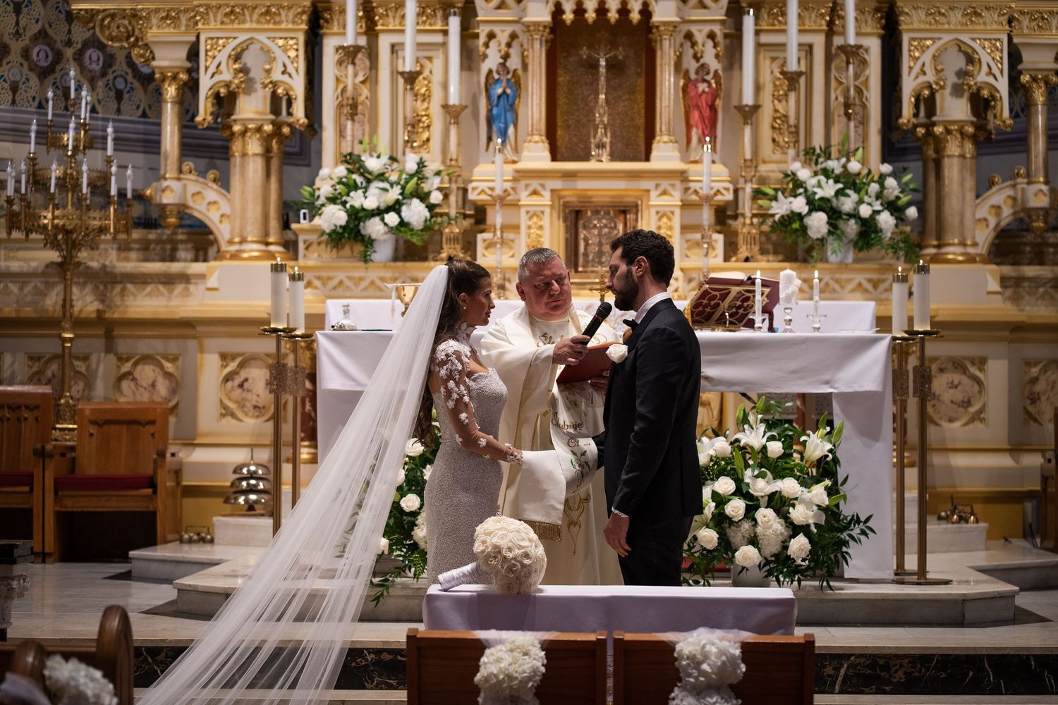 Bride and groom stand before ornate golden altar with white floral arrangements during Catholic wedding ceremony.