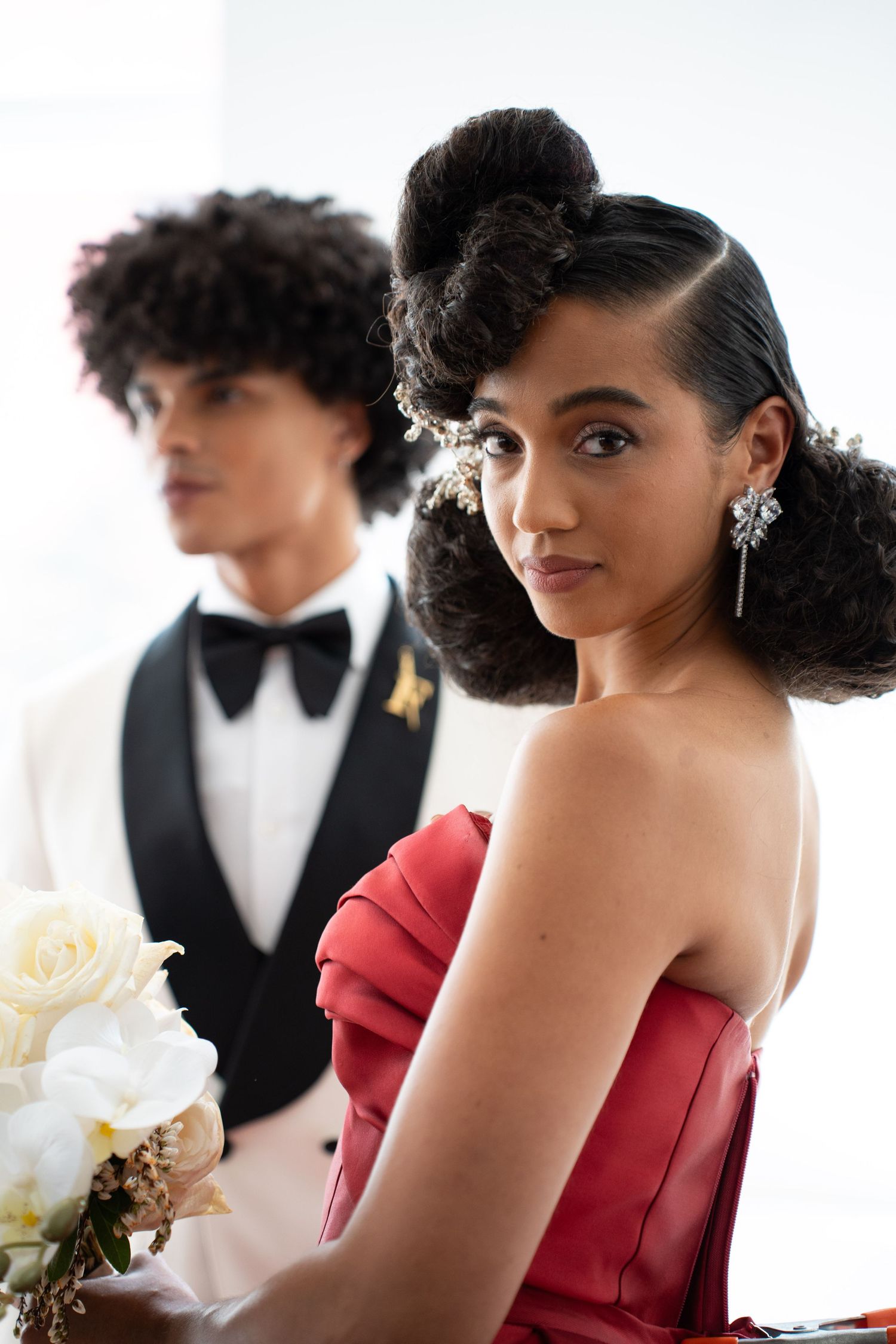 Young woman in red strapless gown with elegant updo and diamond earrings holds white bouquet beside man in tuxedo.