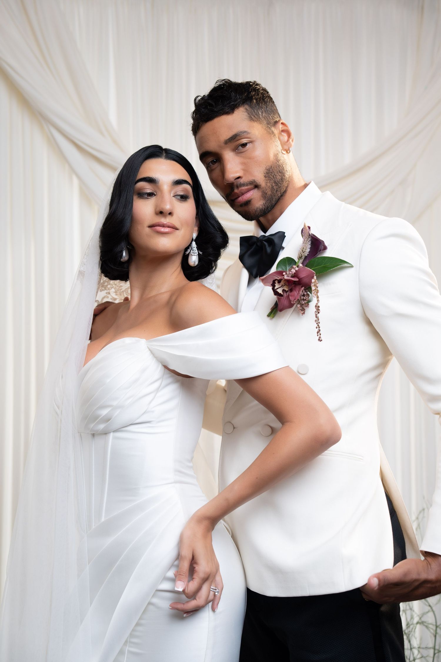 Elegant bride and groom in white wedding outfits pose seriously before ivory draped curtain backdrop.