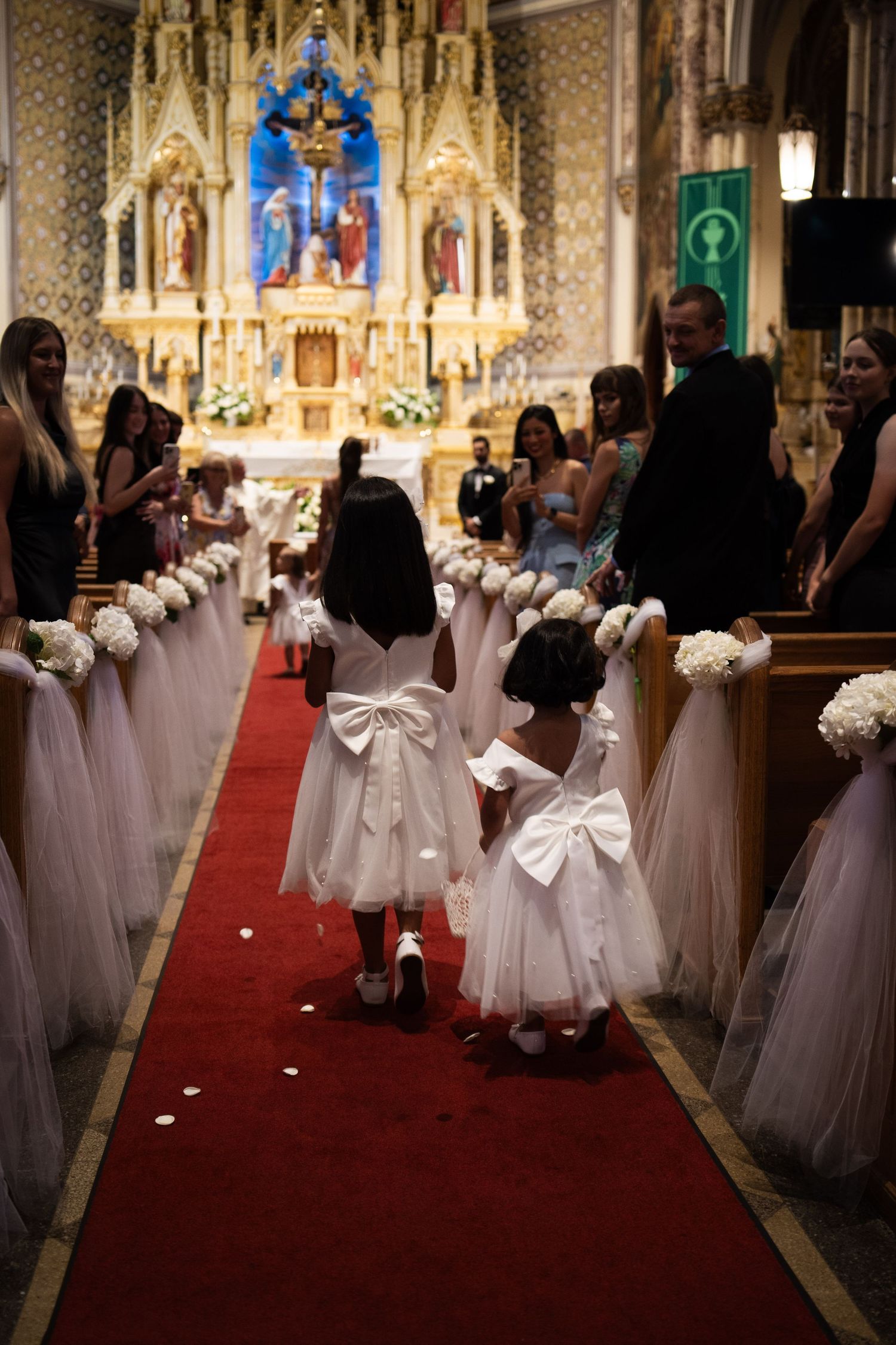 Two young flower girls in white dresses with bows walk down red carpet aisle in ornate church.
