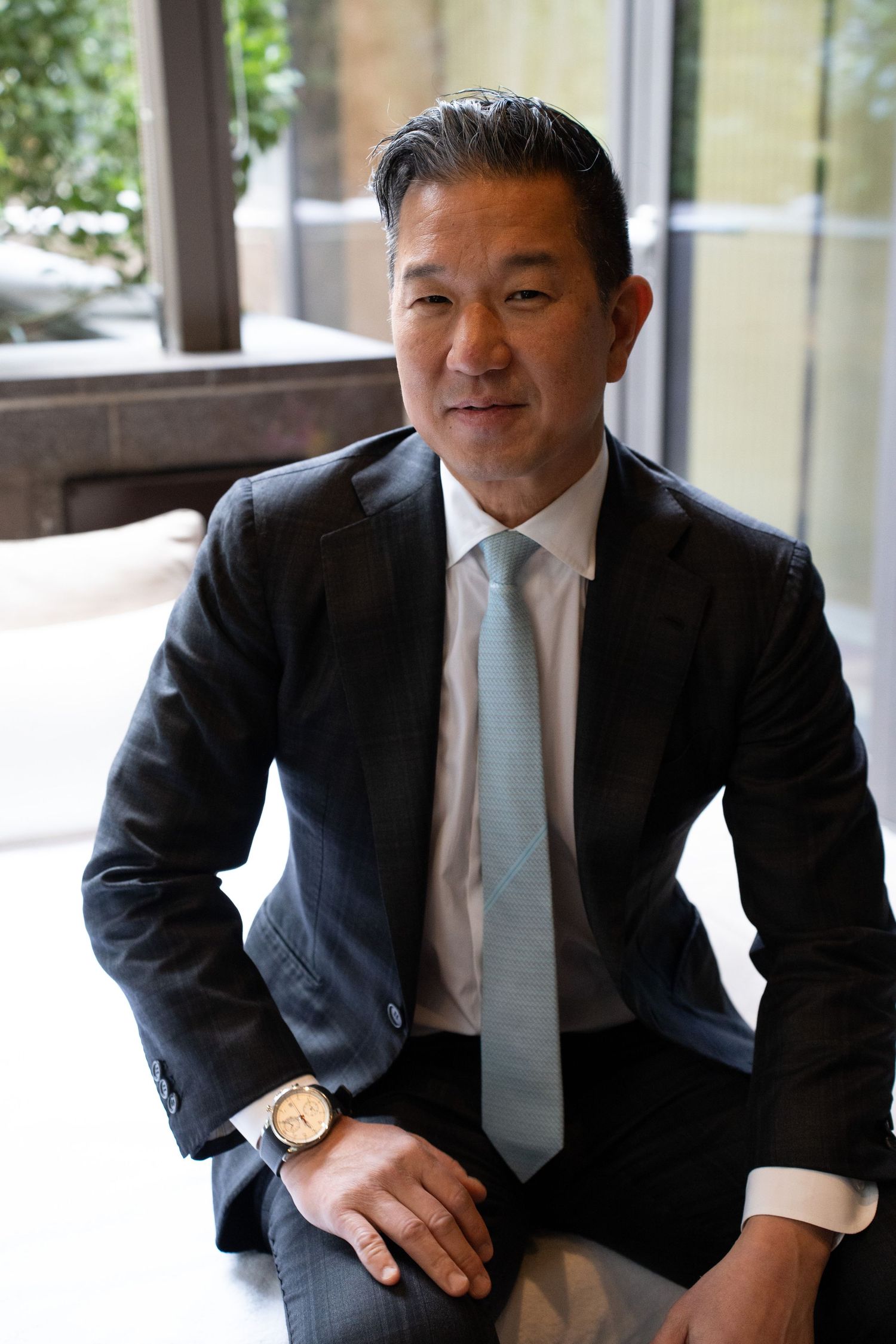 Professional Asian man in dark suit and light blue tie sitting confidently in a modern interior setting.