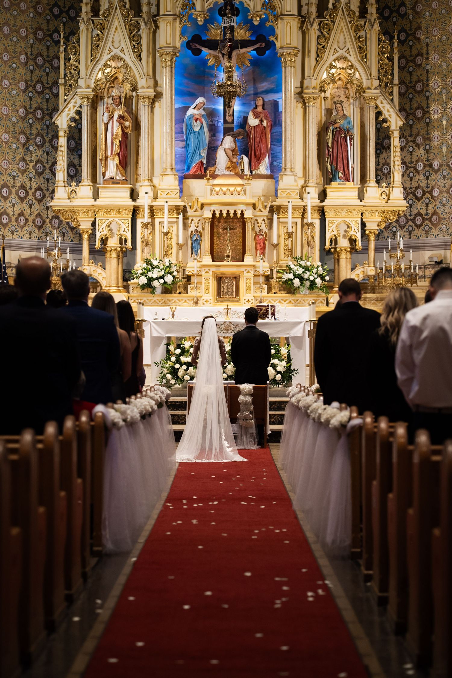 Bride and groom kneeling at altar during church wedding ceremony with red carpet aisle and floral decor.