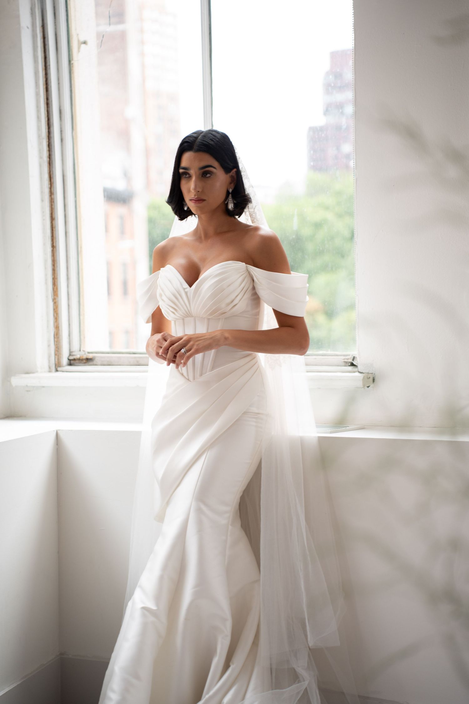 Bride in structured off-shoulder white wedding dress with tulle skirt posing by bright window.