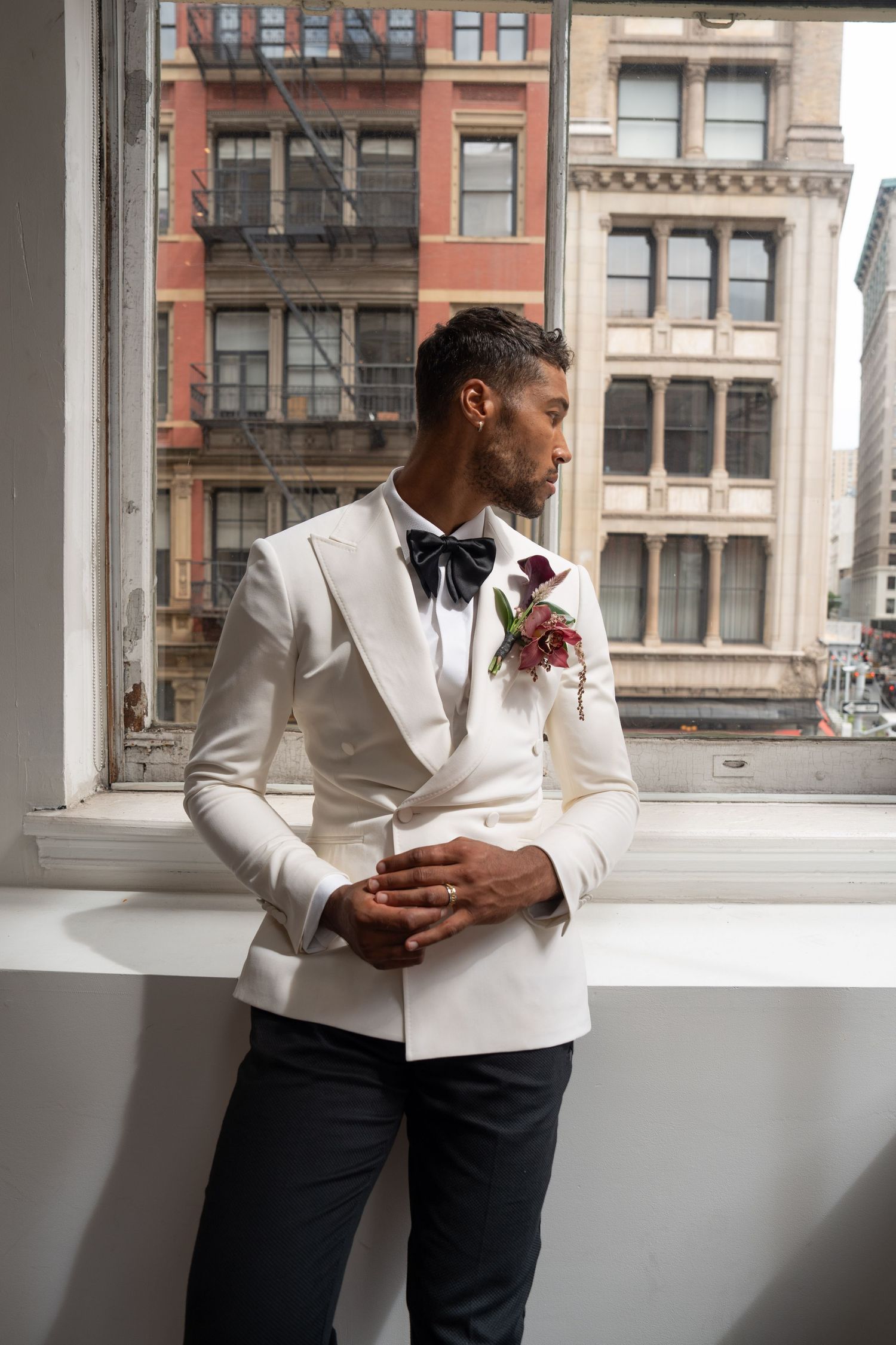 Groom in white double-breasted tuxedo with black bow tie and burgundy boutonniere looks out city window.