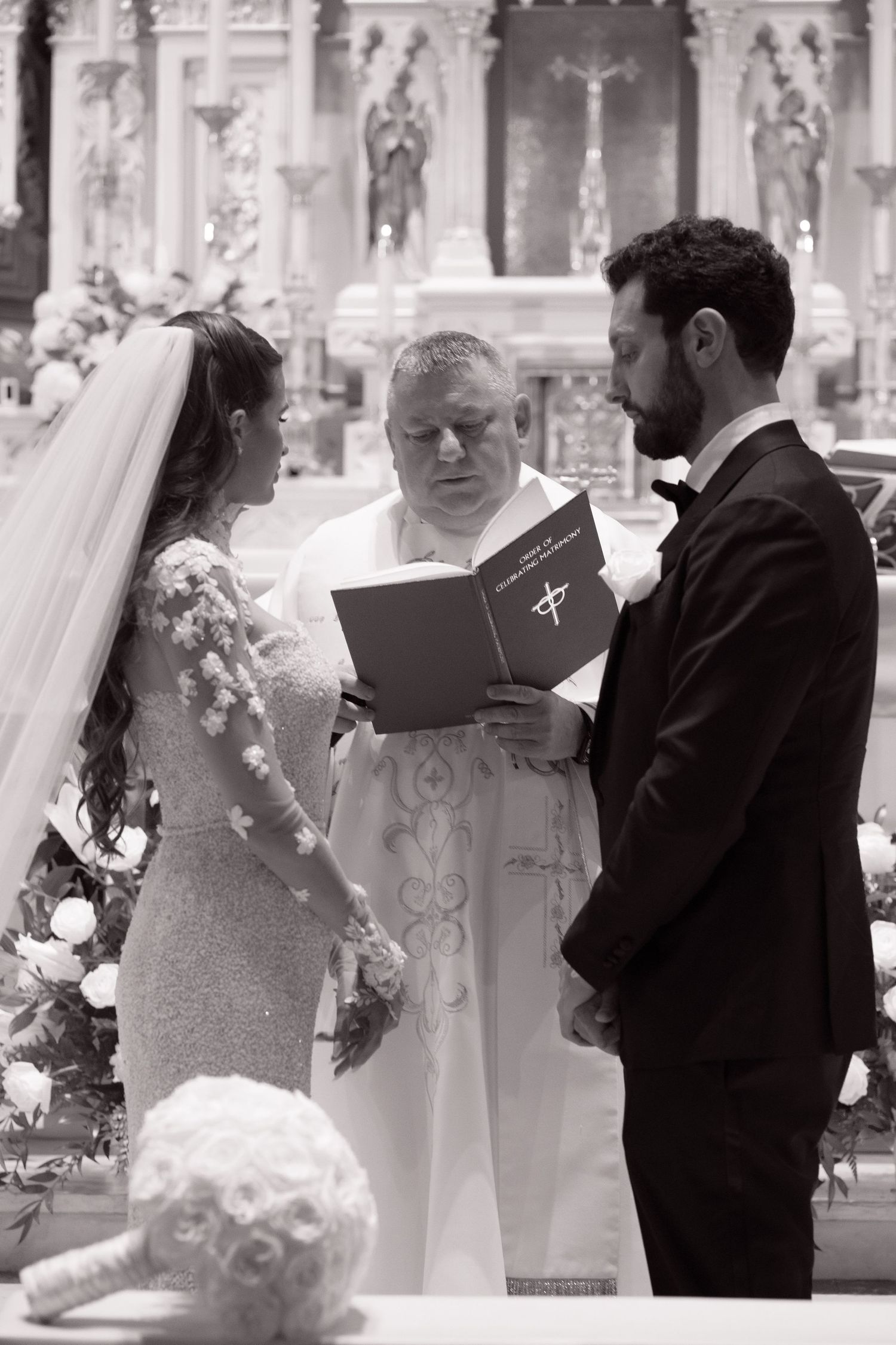 Black and white photo of priest reading vows to bride and groom during Catholic wedding ceremony.