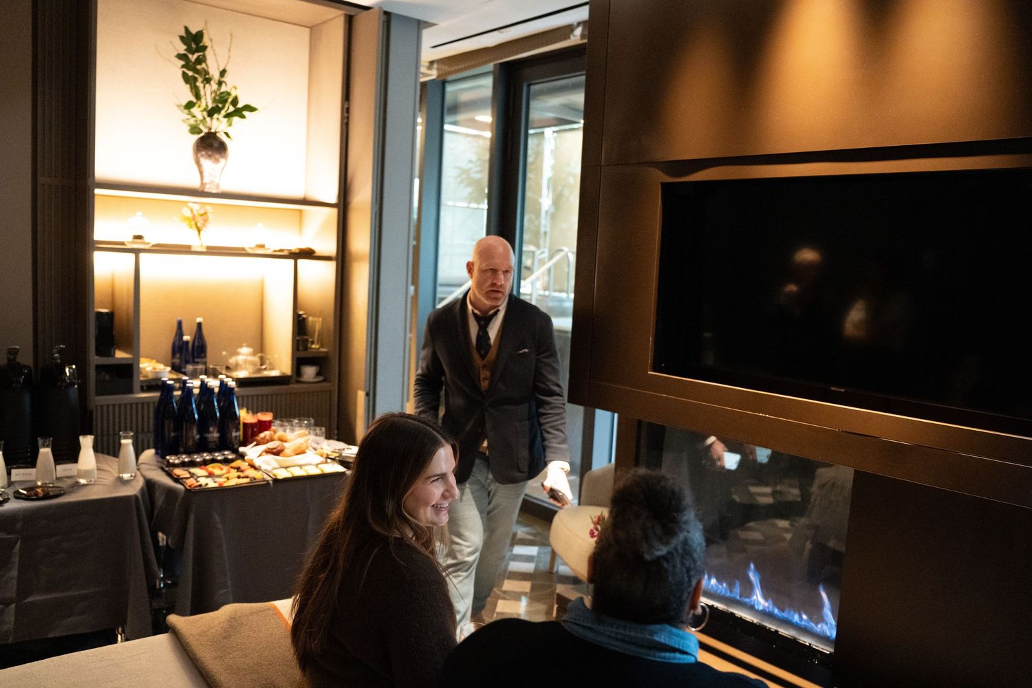 Man in blazer serving guests seated near a modern fireplace at a luxury spa event.