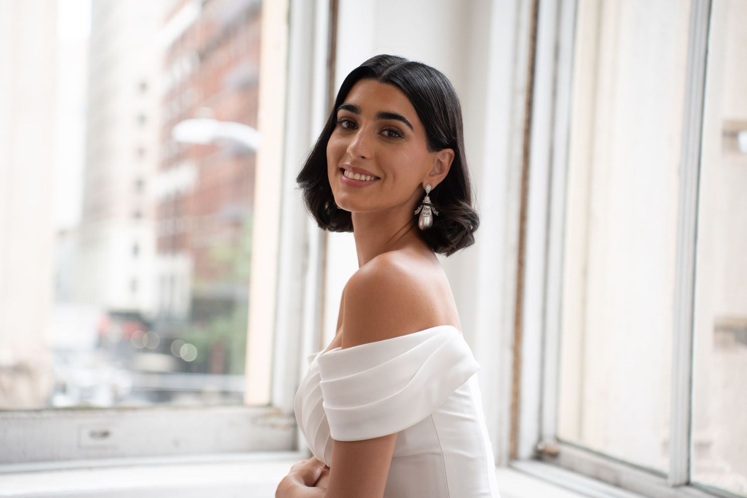 Bride in off-shoulder white gown smiles over shoulder near bright city windows wearing elegant drop earrings.