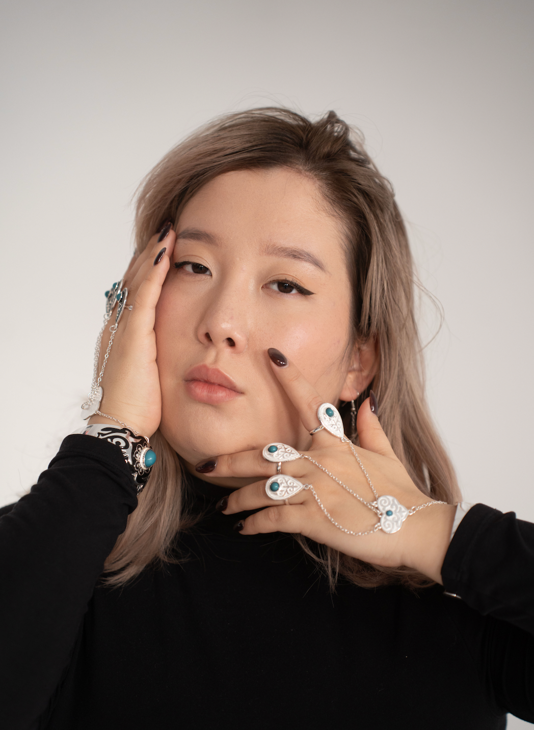 Young woman poses with hands near face wearing silver rings and bracelets against gray background.