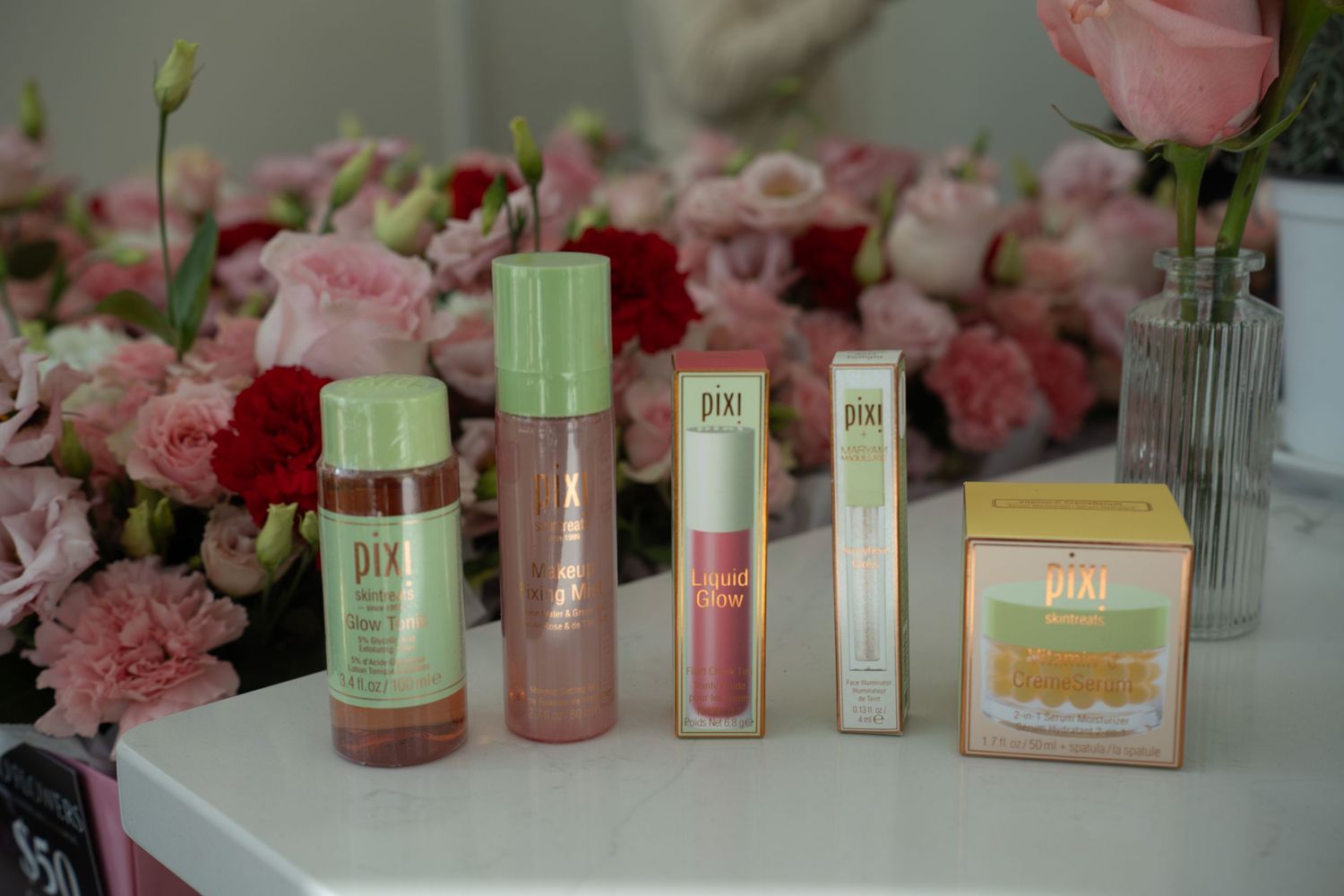 Pixi beauty skincare and makeup products arranged on a white surface with pink flowers behind.