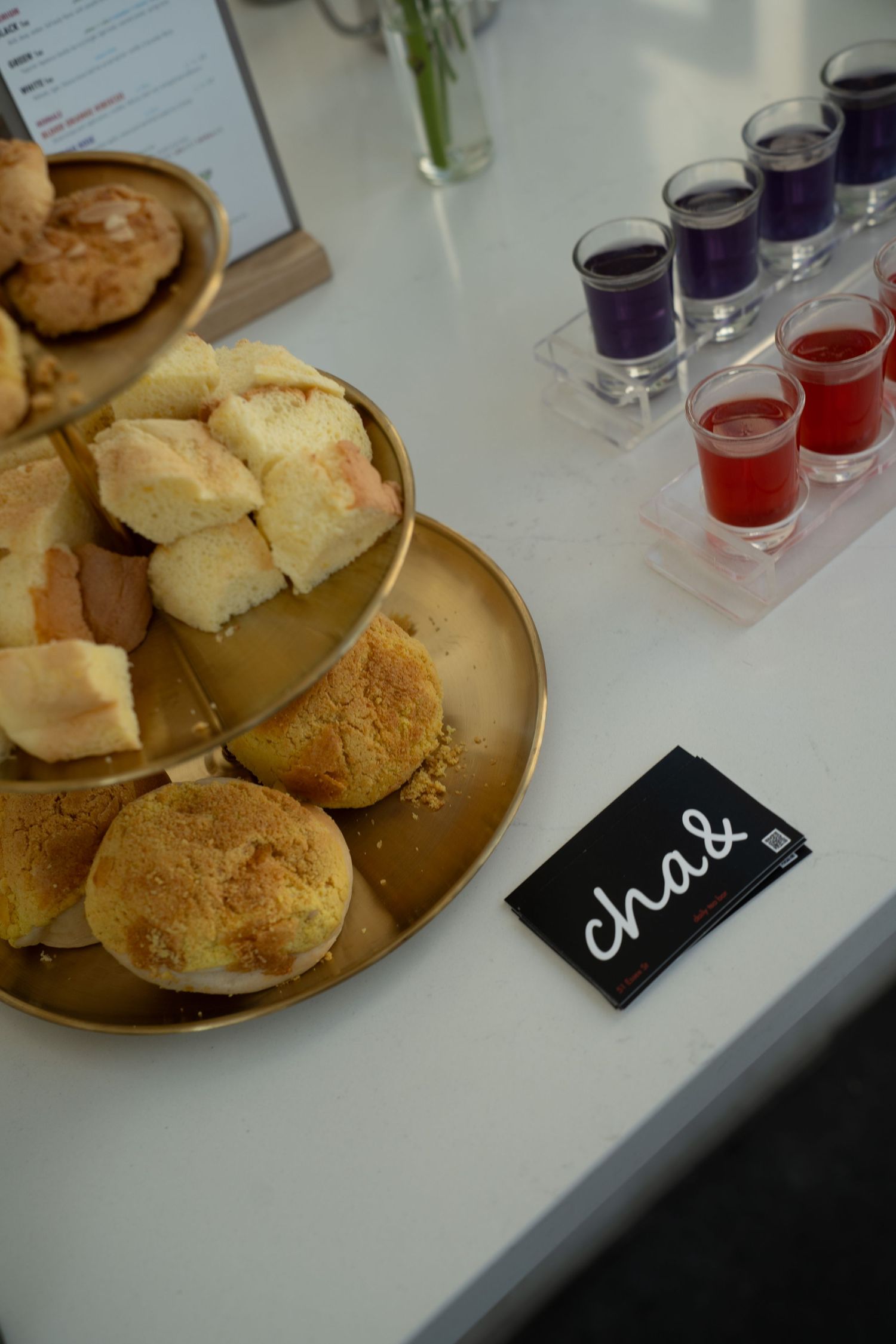 Gold tiered tray with scones beside colorful shot glasses and a cha& card on a white counter.