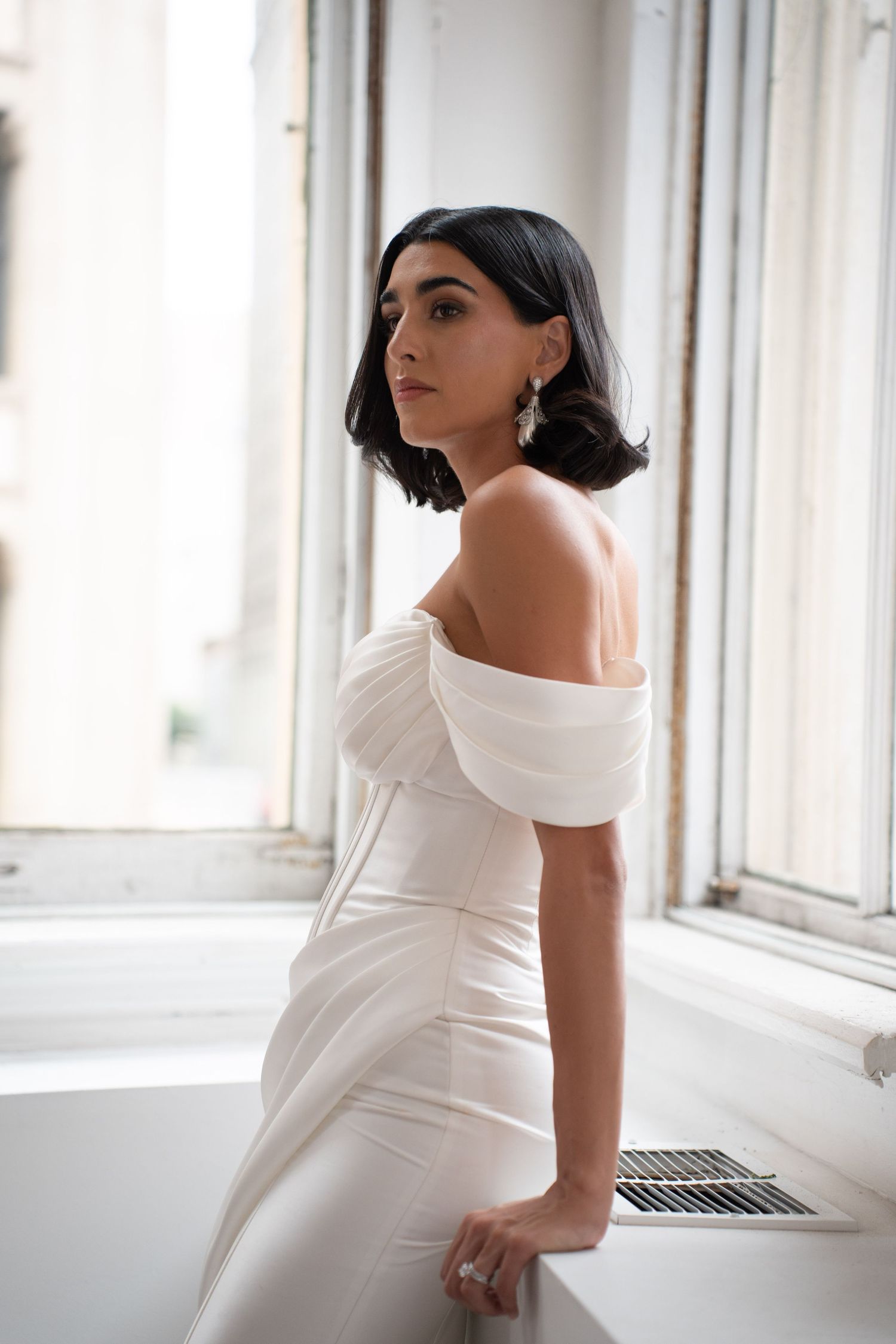Bride in off-shoulder white satin gown gazes over shoulder by bright window in bridal portrait.