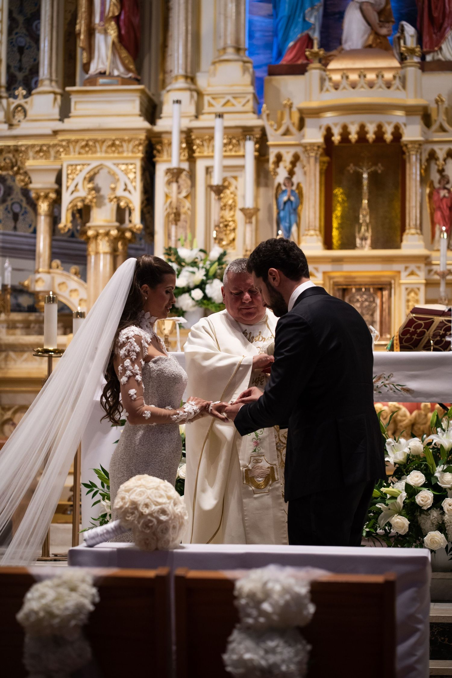 Groom places wedding ring on brides finger as priest oversees ring exchange at decorated church altar.