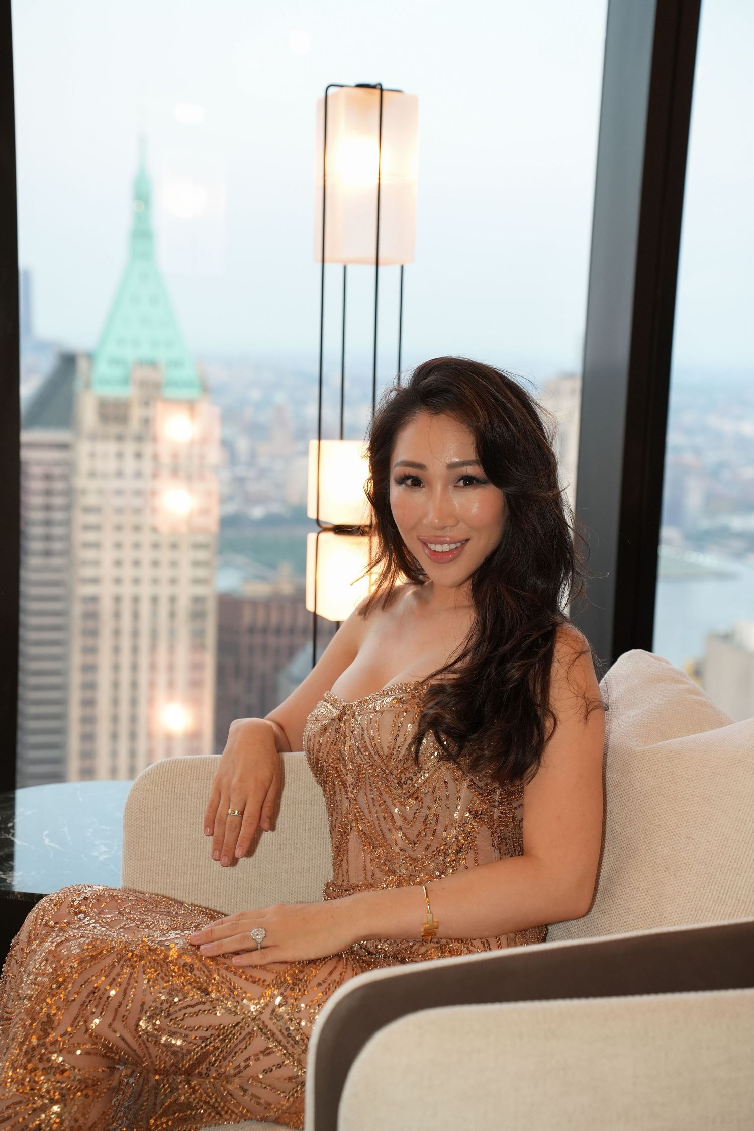 Elegant woman in gold sequin gown seated in luxury high-rise with New York City skyline backdrop.