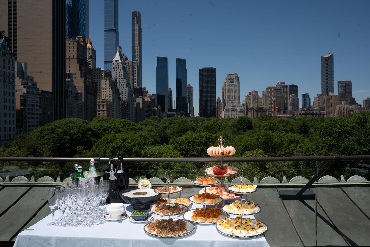 Elegant outdoor brunch spread with tiered pastry display and glassware on terrace overlooking Central Park and NYC skyline.