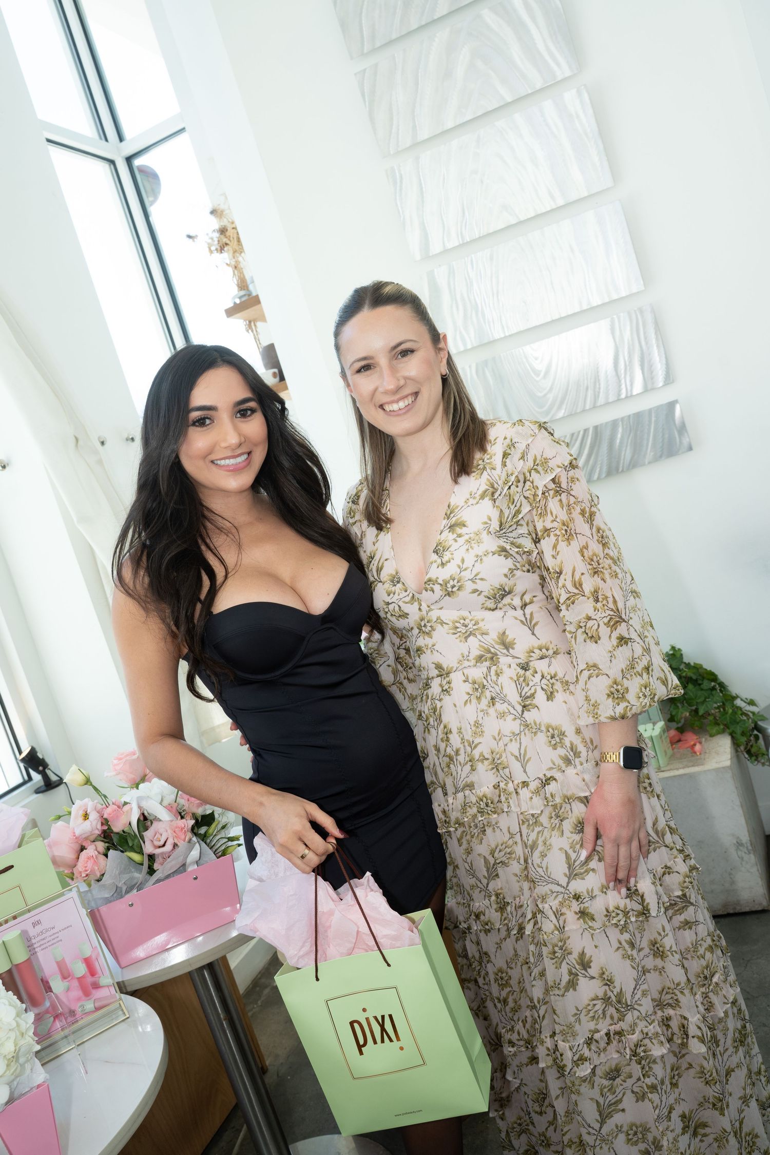 Two women pose with a Pixi beauty gift bag beside a floral arrangement at a brand event.