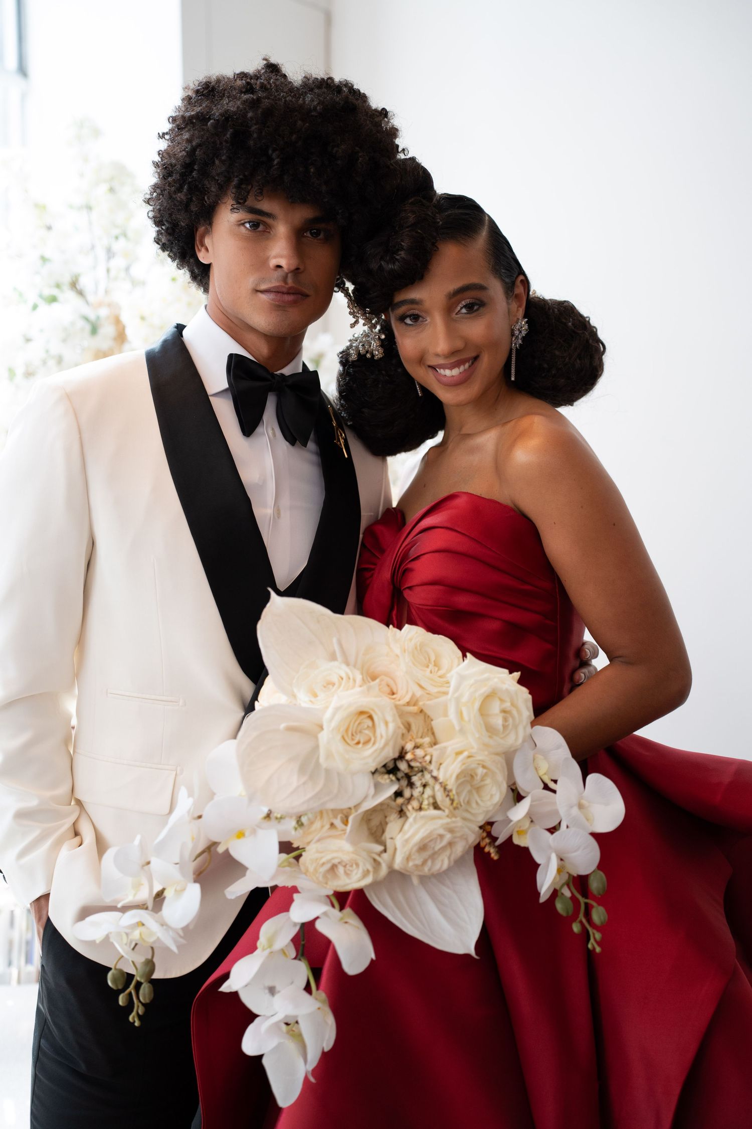 Stylish wedding couple smiling, groom in white tuxedo, bride in red satin gown holding cascading white orchid and rose bouquet.