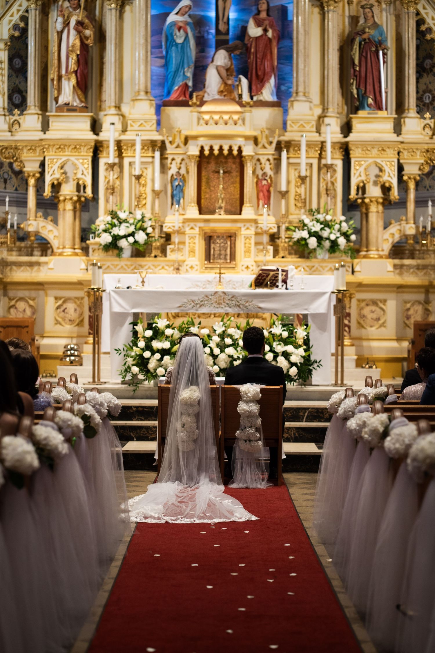 Bride and groom kneeling before elaborately decorated golden Catholic church altar with white floral arrangements.