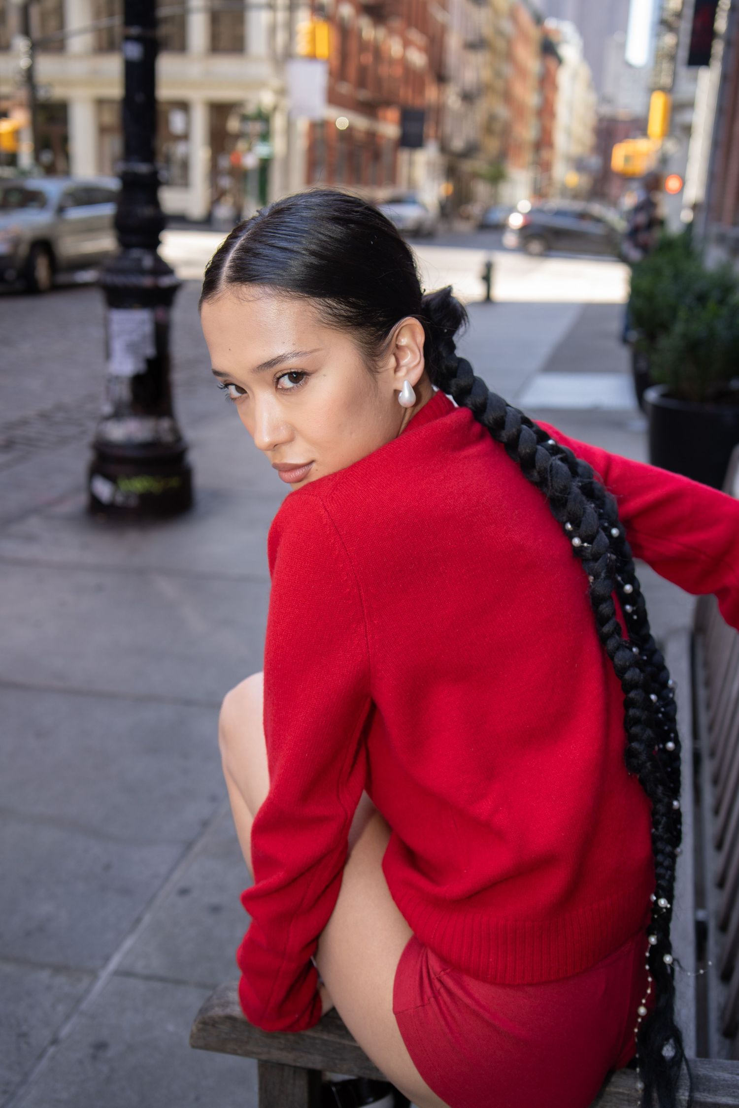 Woman in red sweater and skirt with long braided hair poses on a bench on a city street.