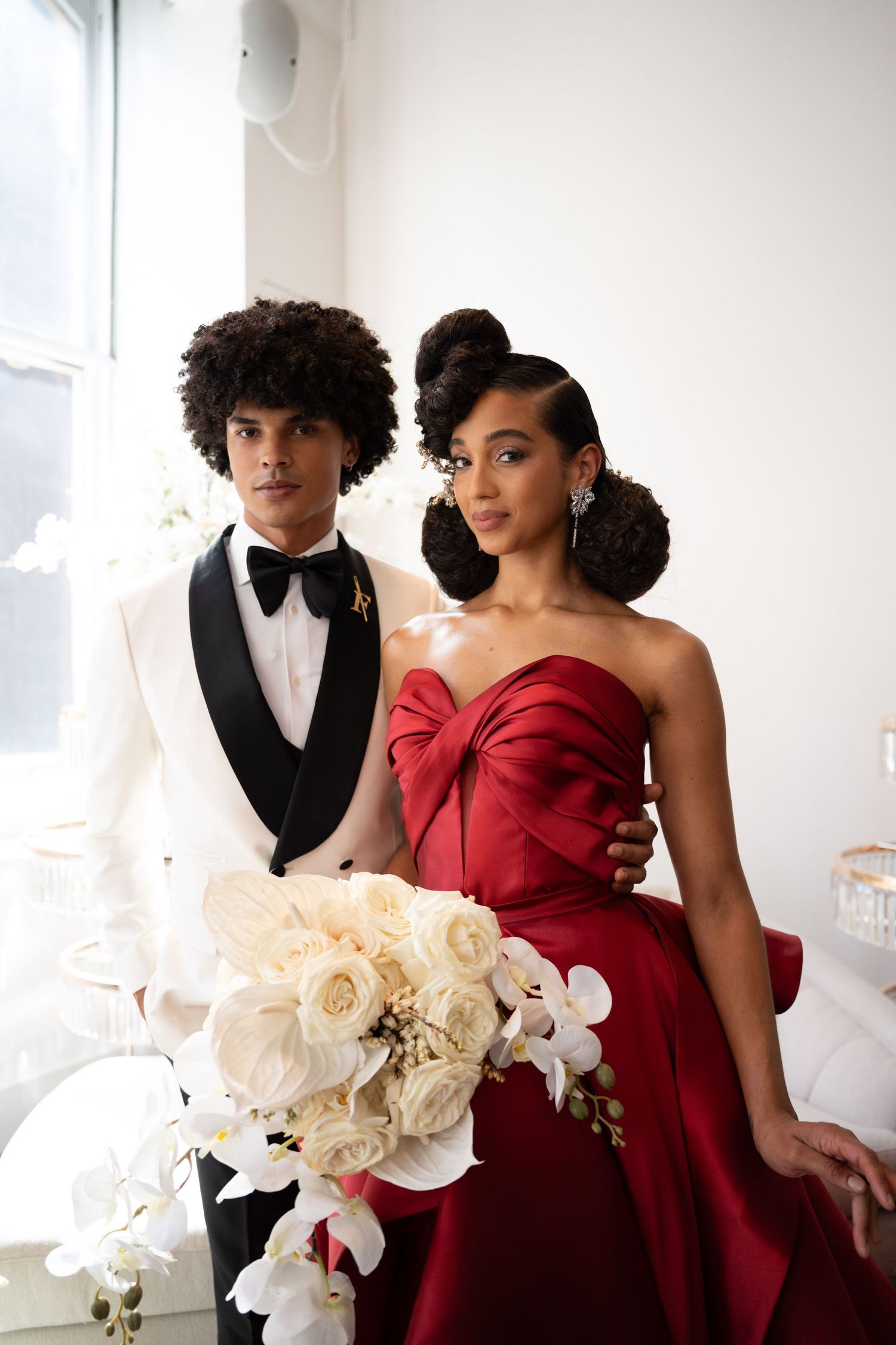 Wedding couple in red satin gown and white tuxedo posing with cascading white rose bouquet.