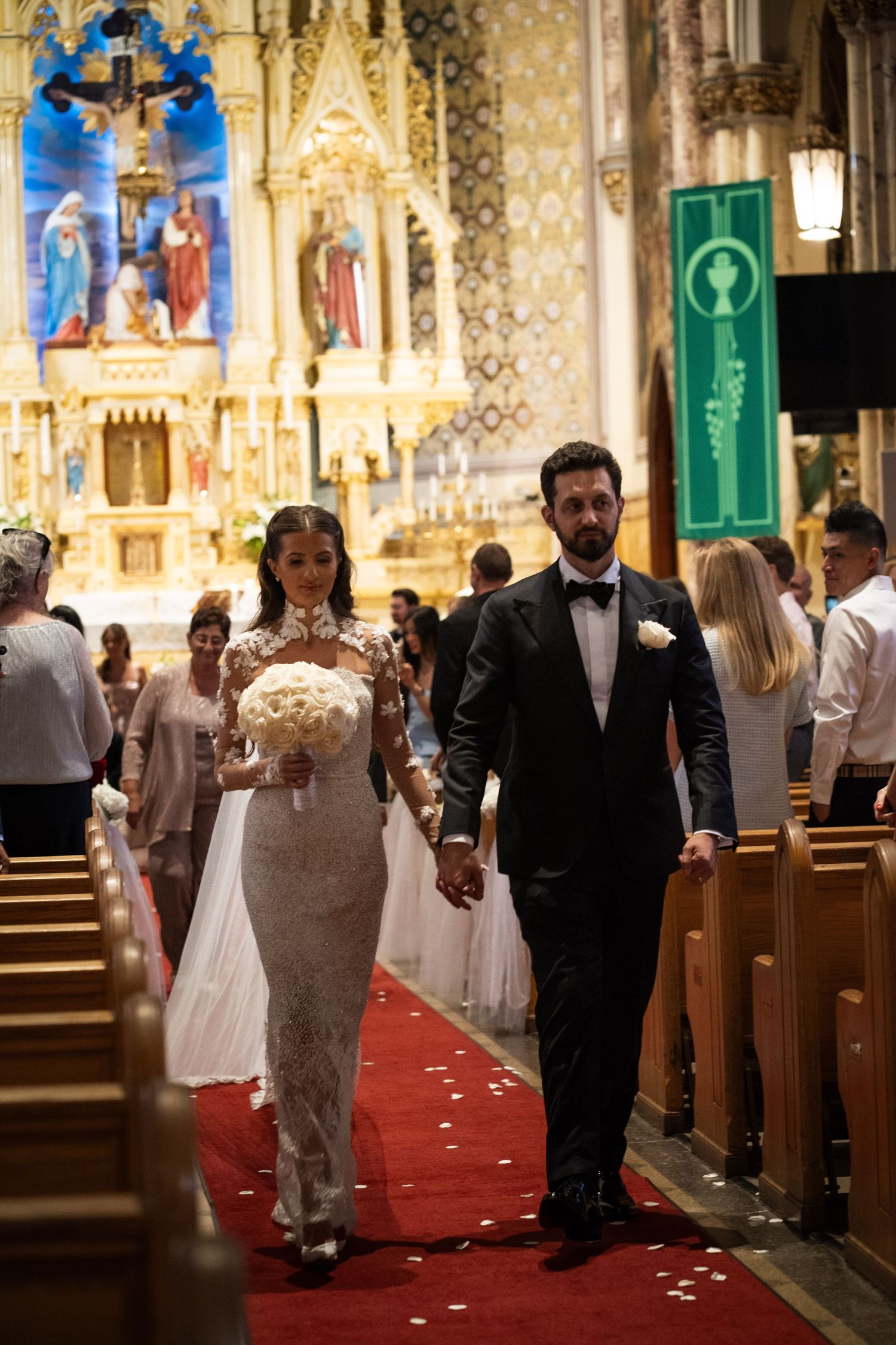 Bride and groom walk down red carpet aisle of ornate Catholic church after wedding ceremony.