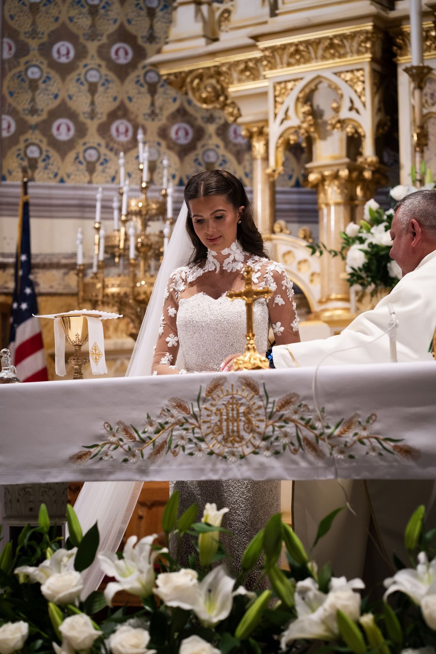 Bride in lace wedding gown participates in Catholic Mass at elaborately decorated church altar.