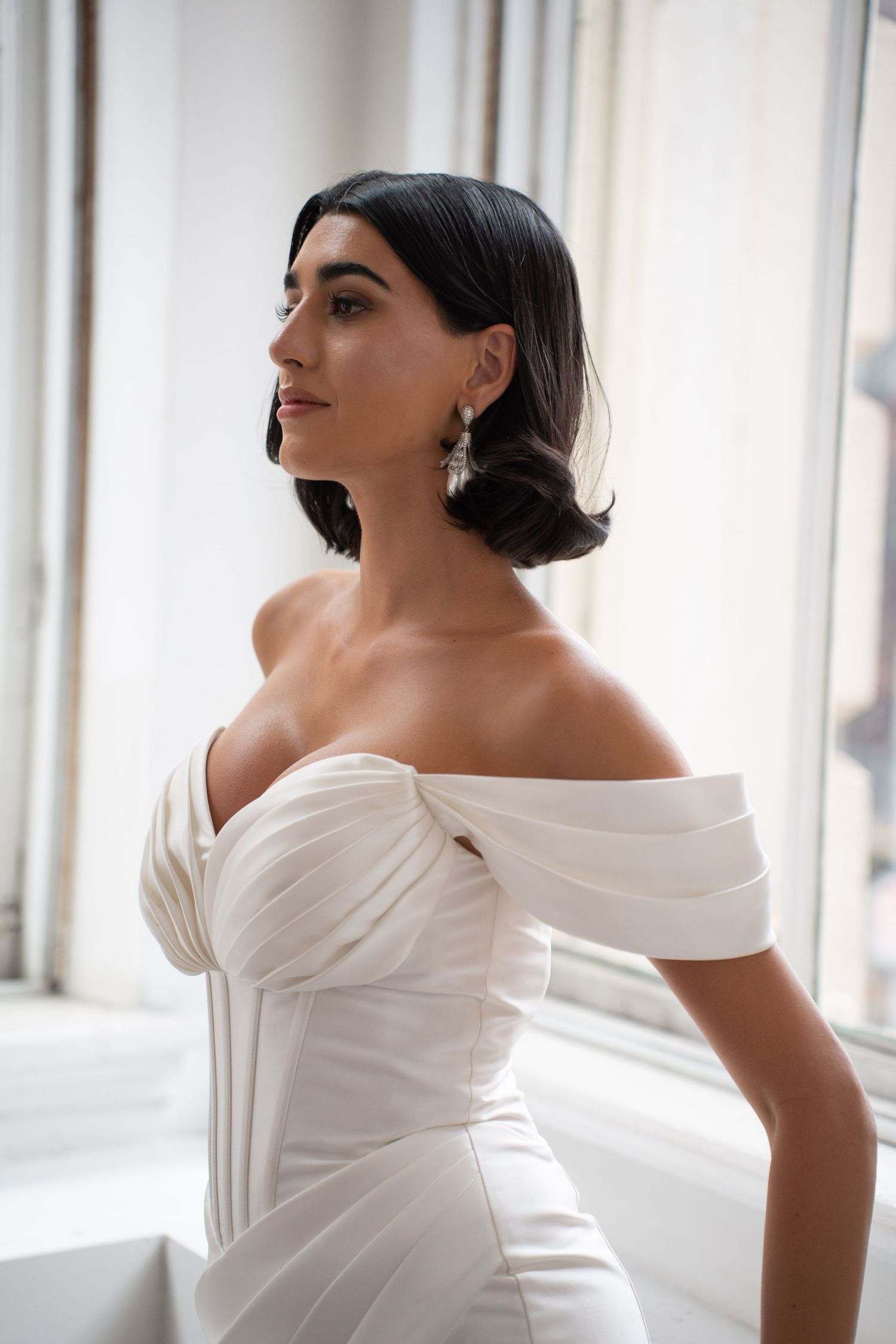 Elegant bride in structured off-shoulder white corset wedding gown with short dark hair gazes toward window light.
