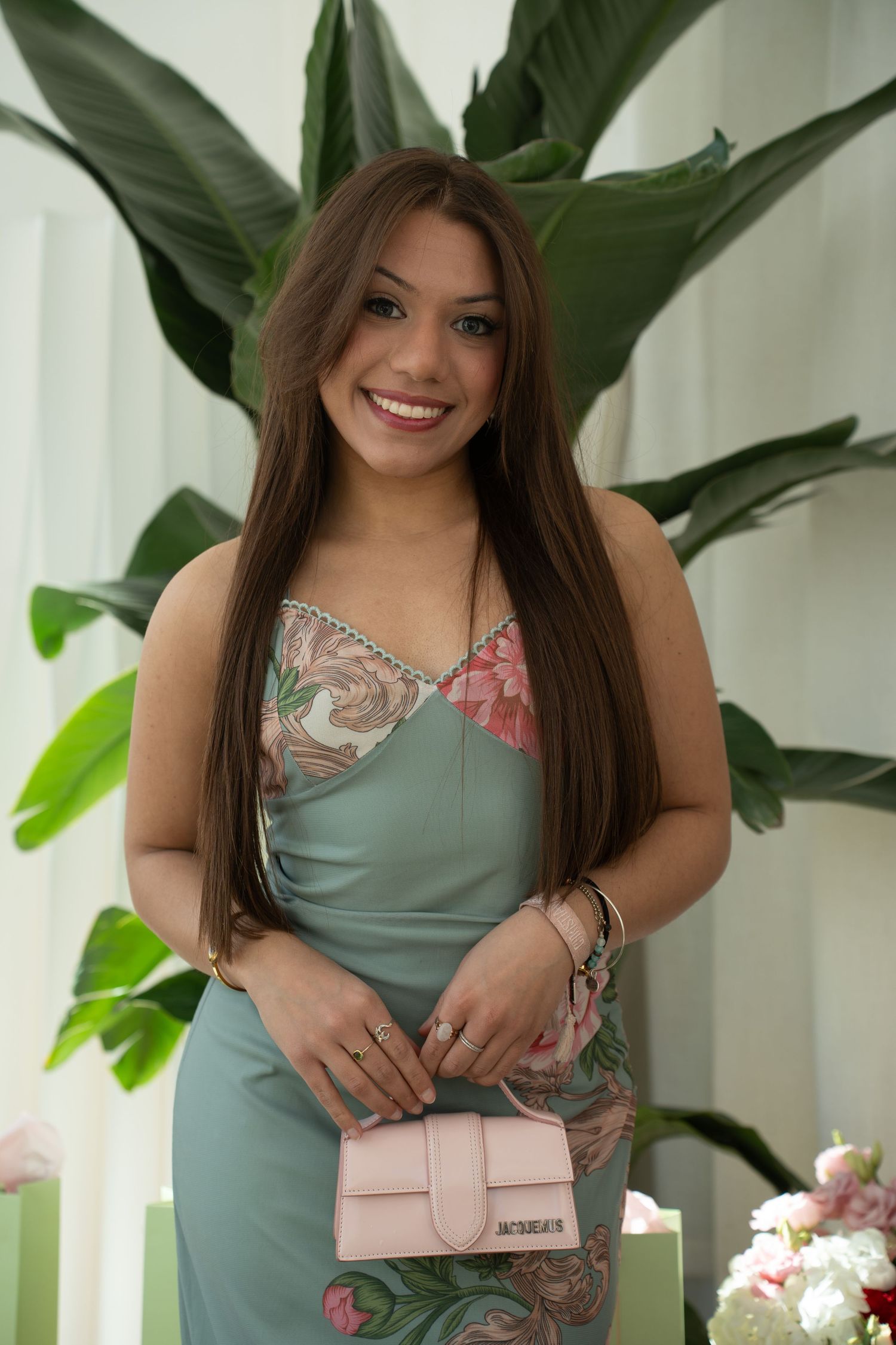 Smiling woman in green floral dress holding pink Jacquemus bag with tropical plants behind her.