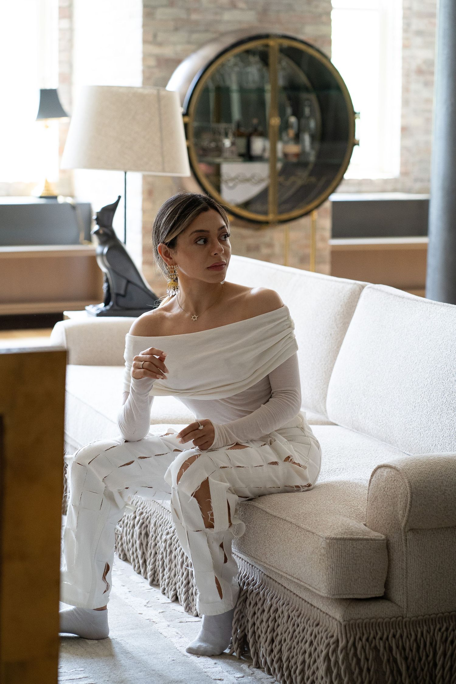 Woman in white off-shoulder outfit sitting on cream sofa in elegant interior setting.