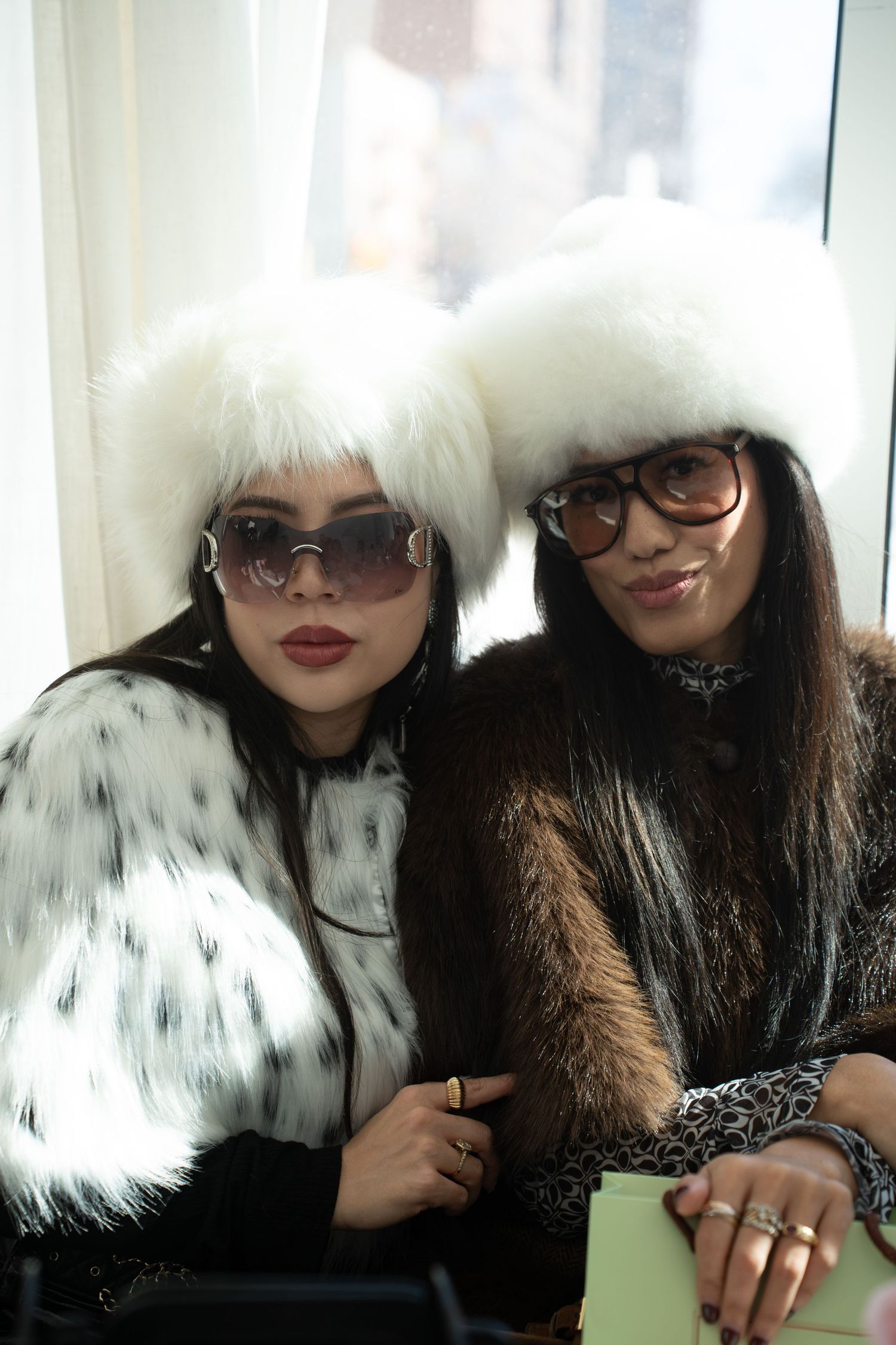 Two women pose in matching white fur hats and fur coats wearing stylish sunglasses.