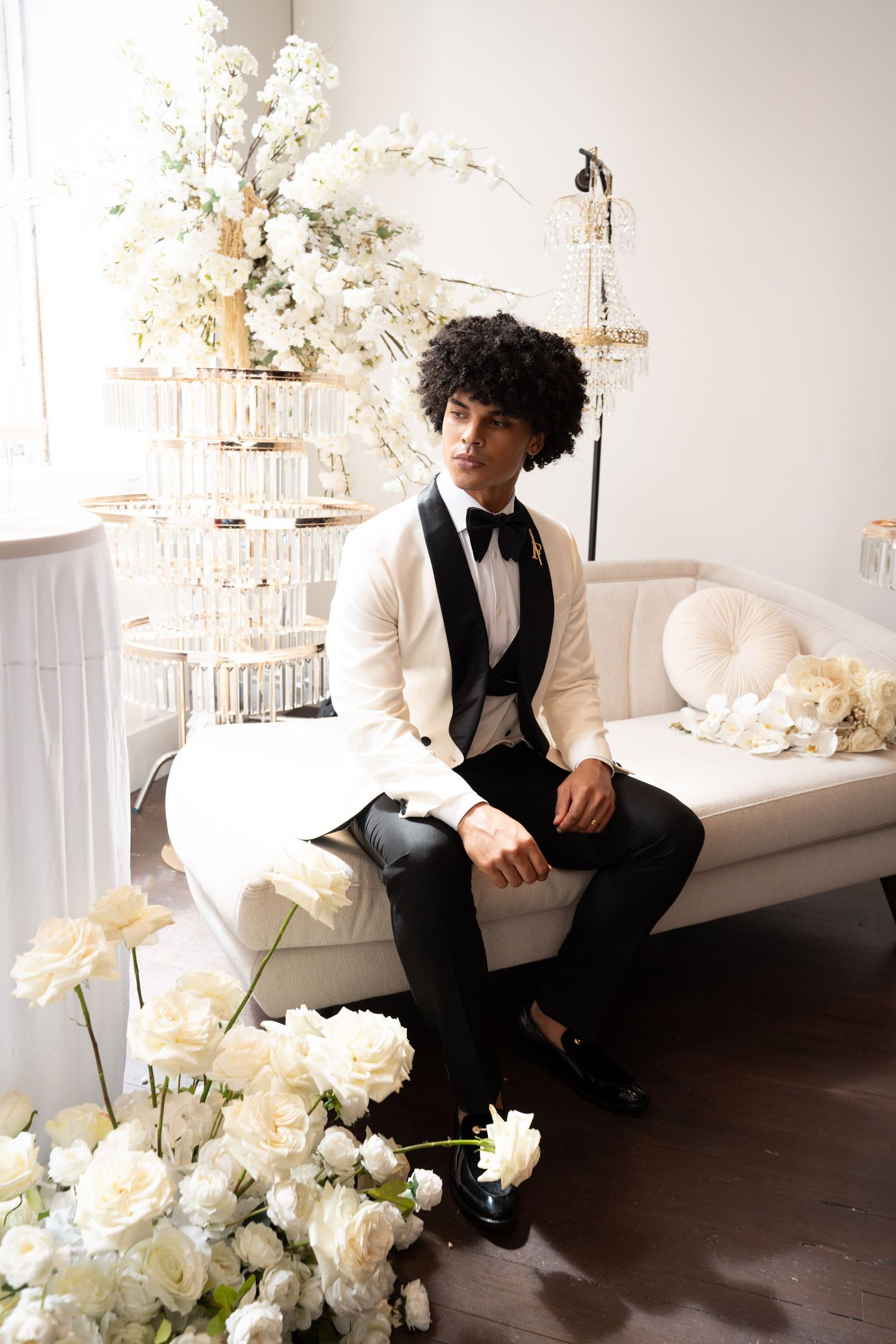 Elegant groom in white tuxedo sits on ivory sofa surrounded by white floral arrangements at wedding.