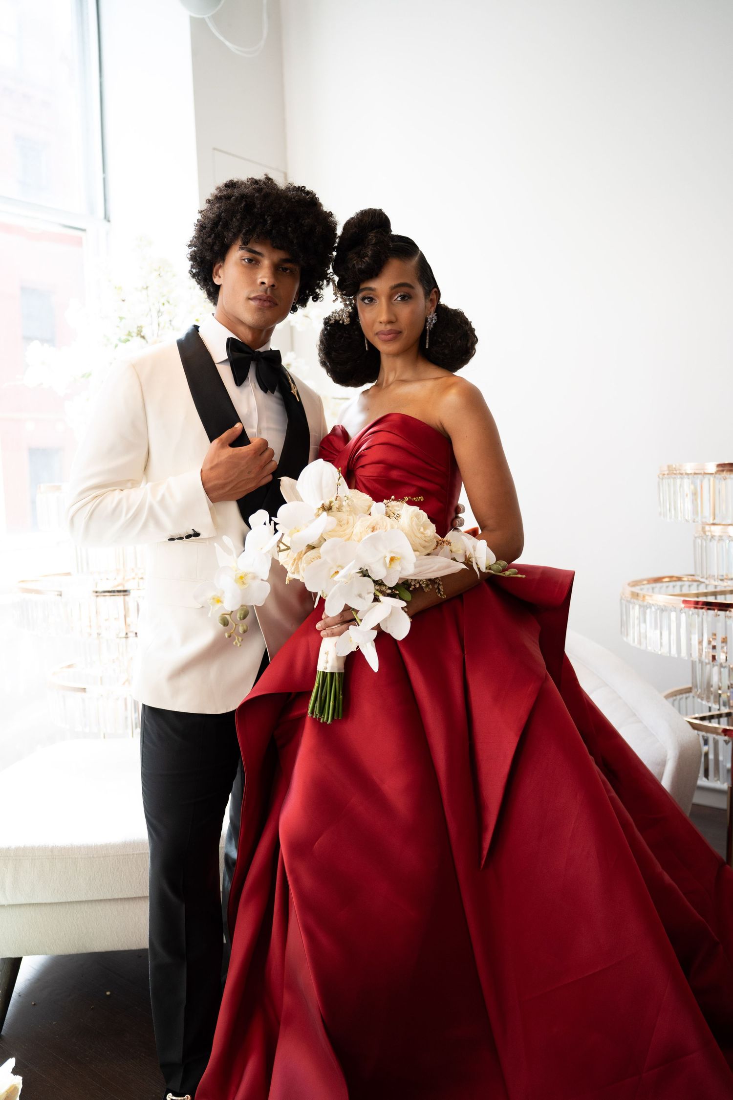 Black wedding couple posing together, groom in white tuxedo, bride in red ball gown with white orchid bouquet.