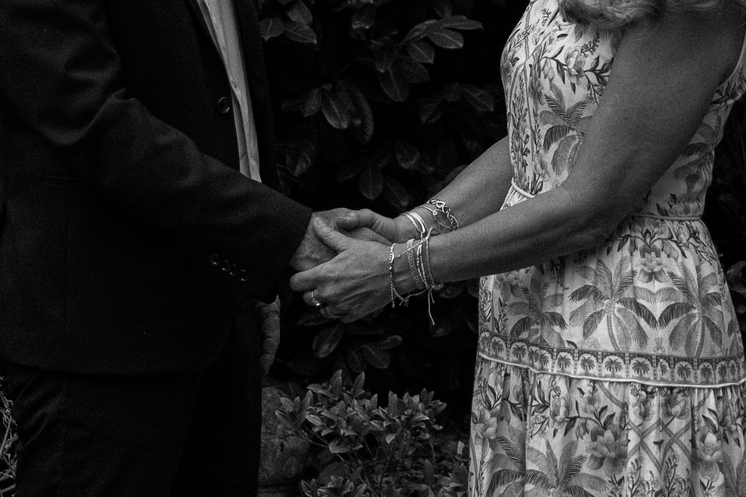 A black and white photo showing two people holding hands during a wedding ceremony with intricate dress details visible.
