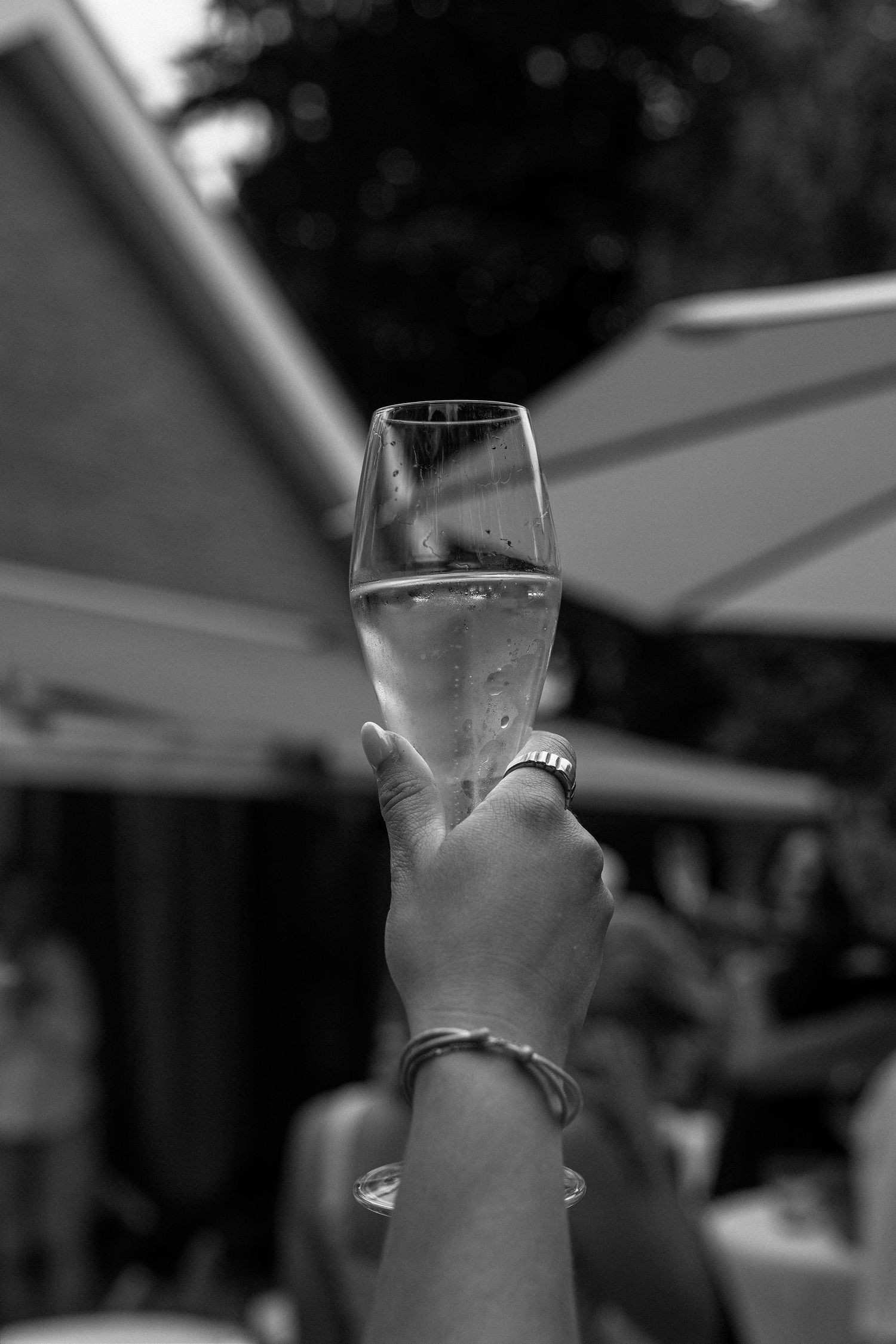 A hand holding up a champagne flute with bubbling liquid against a blurred outdoor background in black and white.