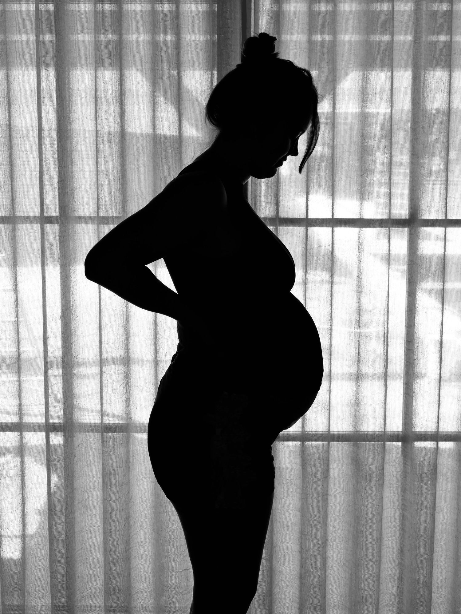 Zwangere vrouw silhouet tegen gordijnraam in zwart-wit.