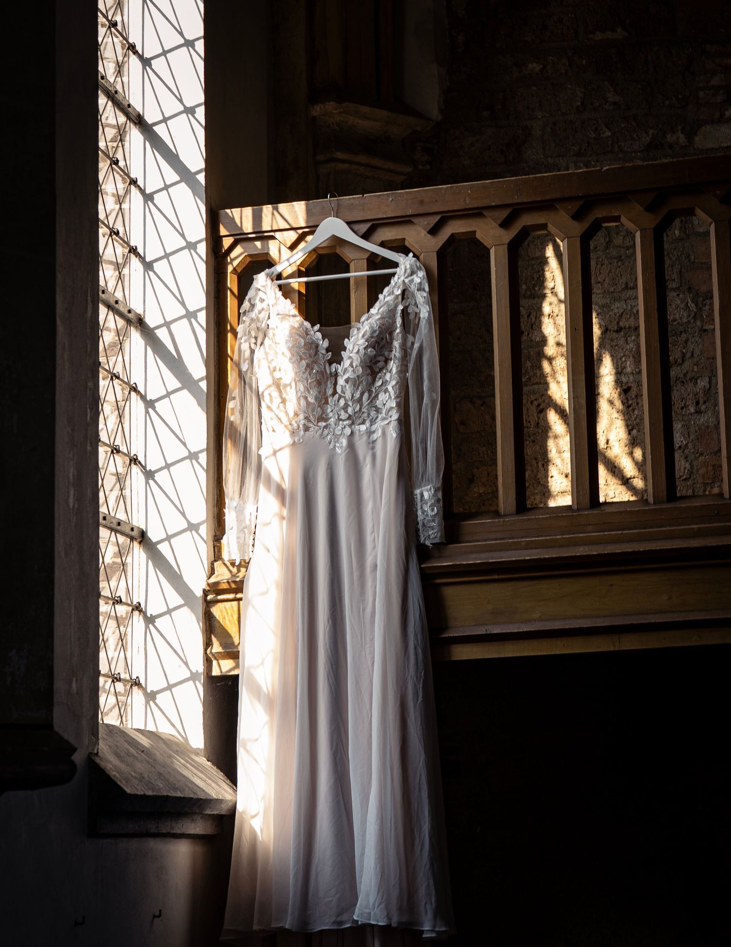 Witte trouwjurk hangend in kerkraam met zonlicht.