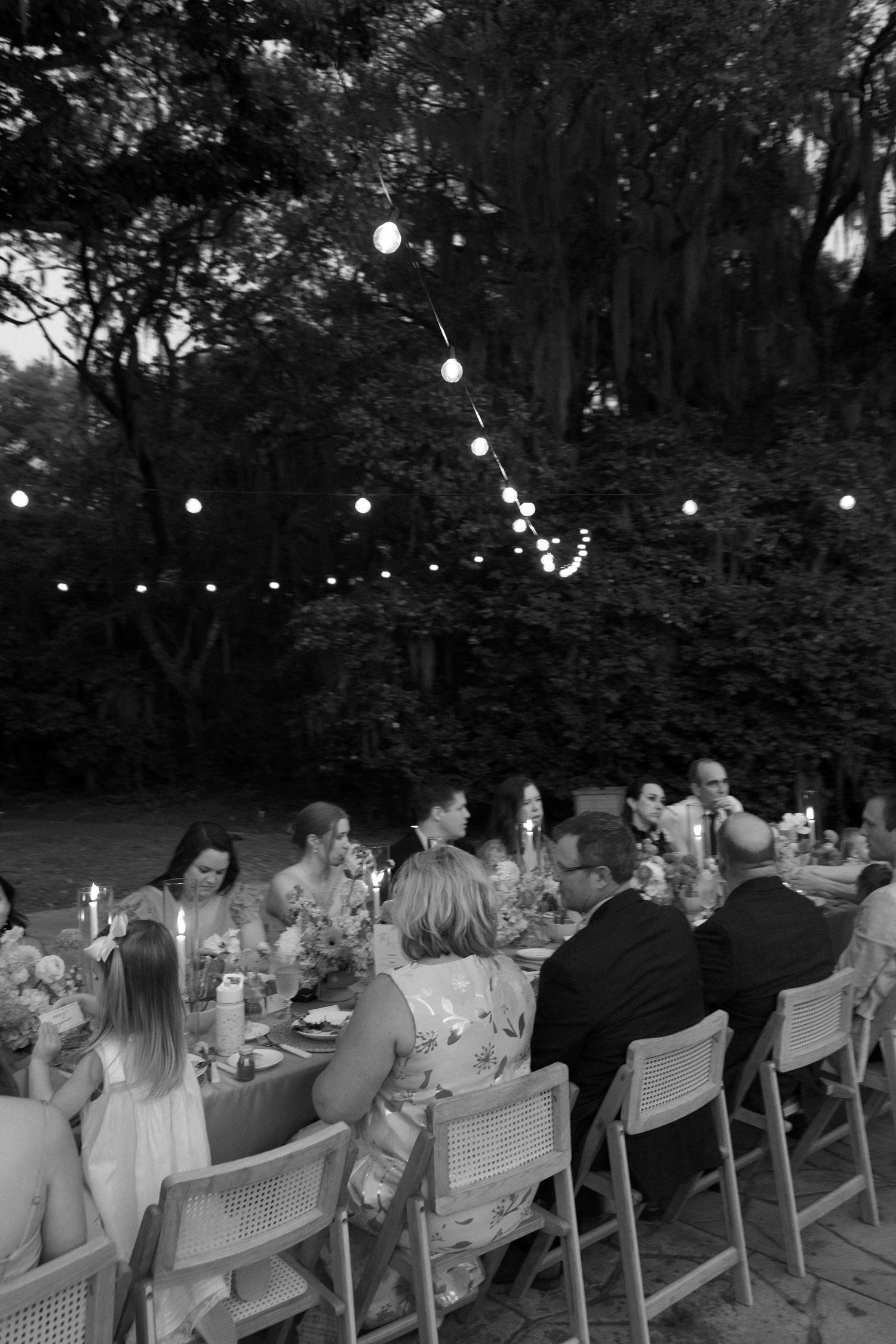 Black and white outdoor evening dinner party with string lights hanging over guests seated at a long decorated table.