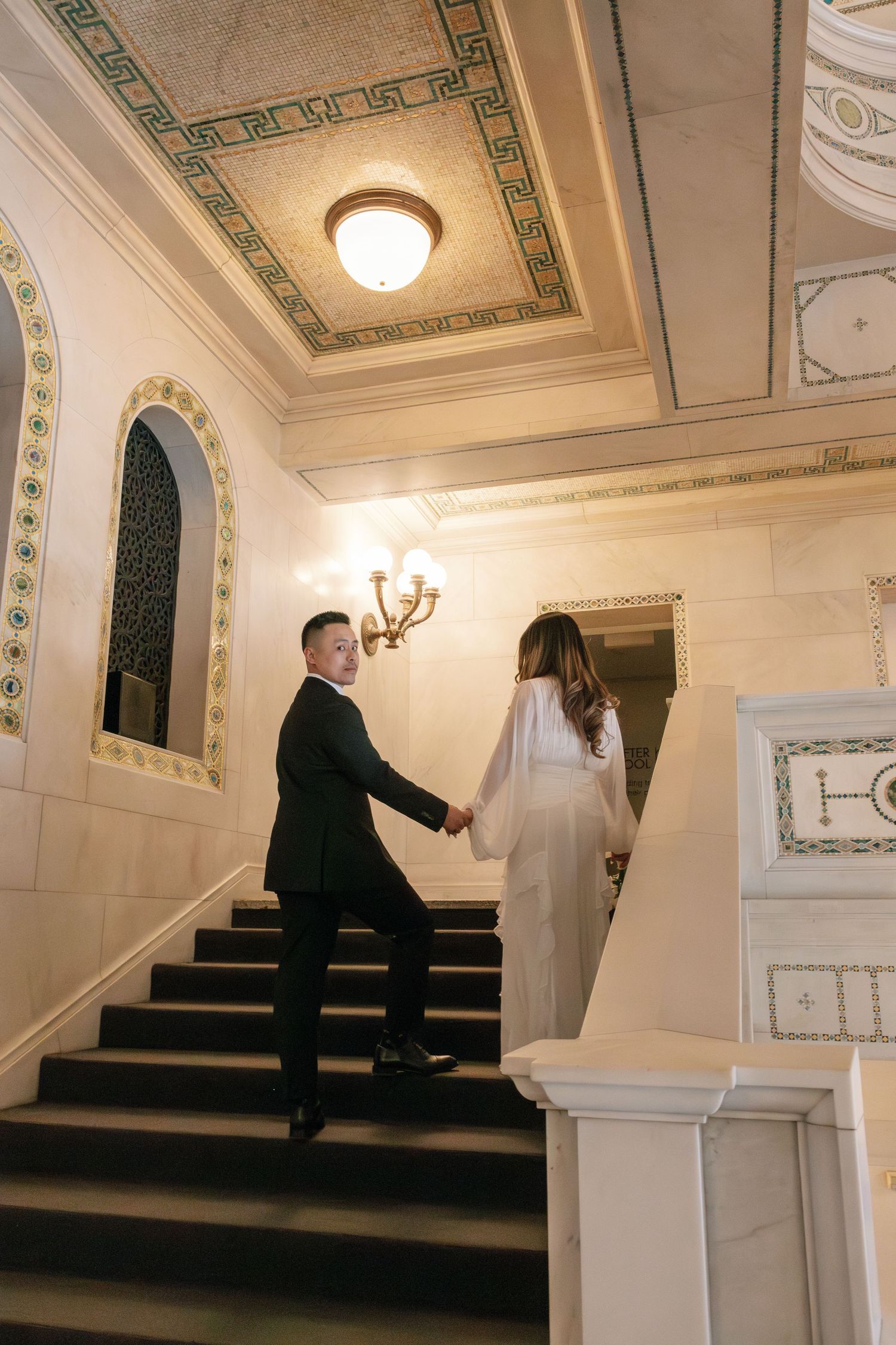 A person in a dark suit ascending an ornate white marble staircase with decorative moldings and ceiling details.