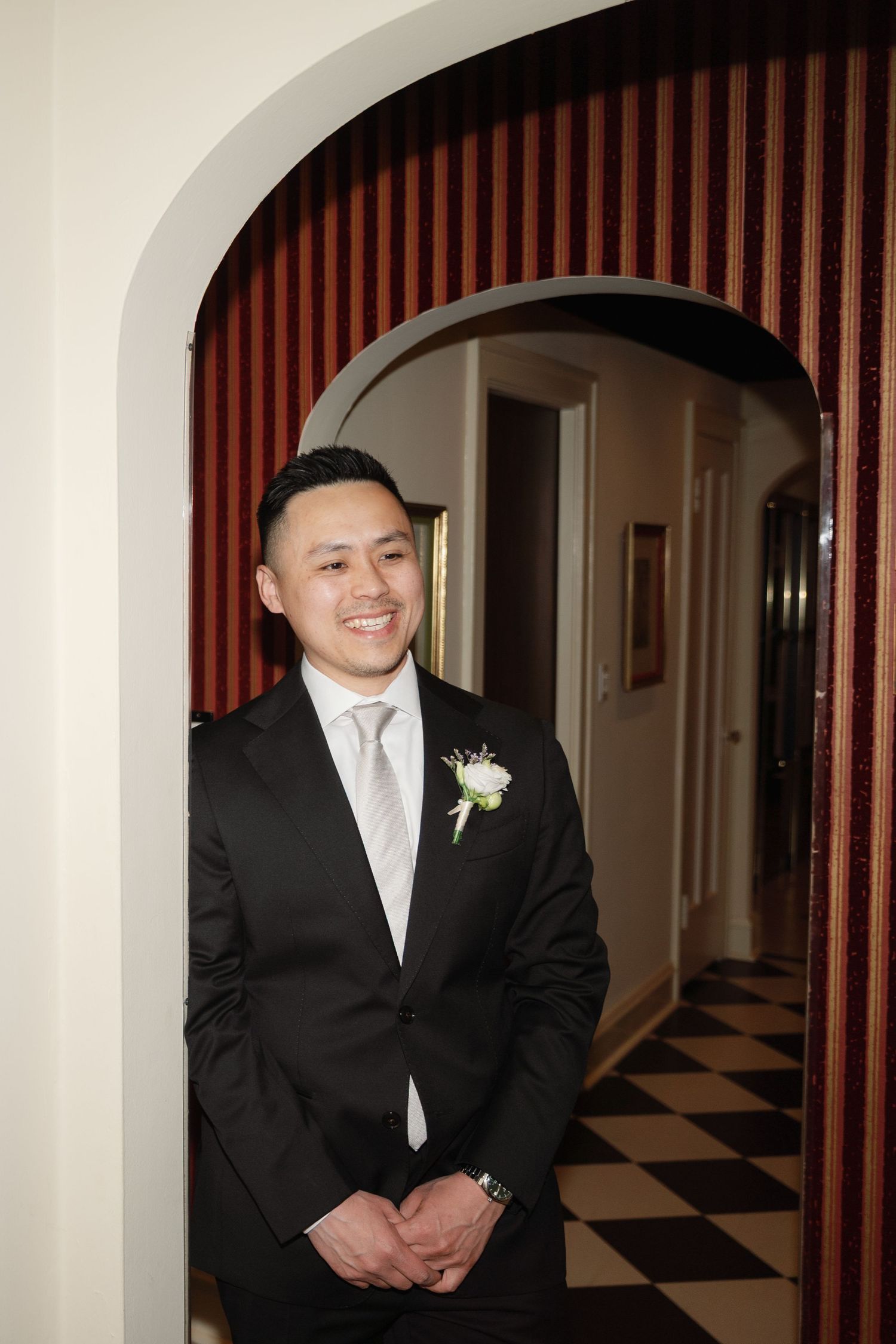 A groom in a black suit and silver tie stands in an arched doorway with a boutonniere on his lapel.