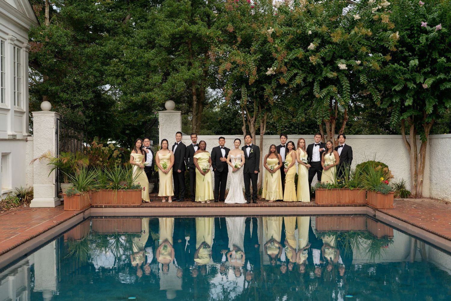 Wedding party stands on stone steps overlooking a blue swimming pool in an elegant garden setting.