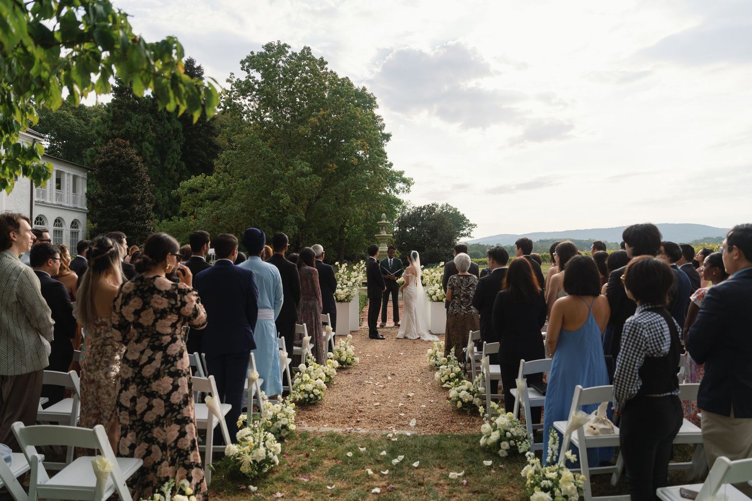 People gather outdoors for a scenic wedding ceremony overlooking a natural landscape at sunset.