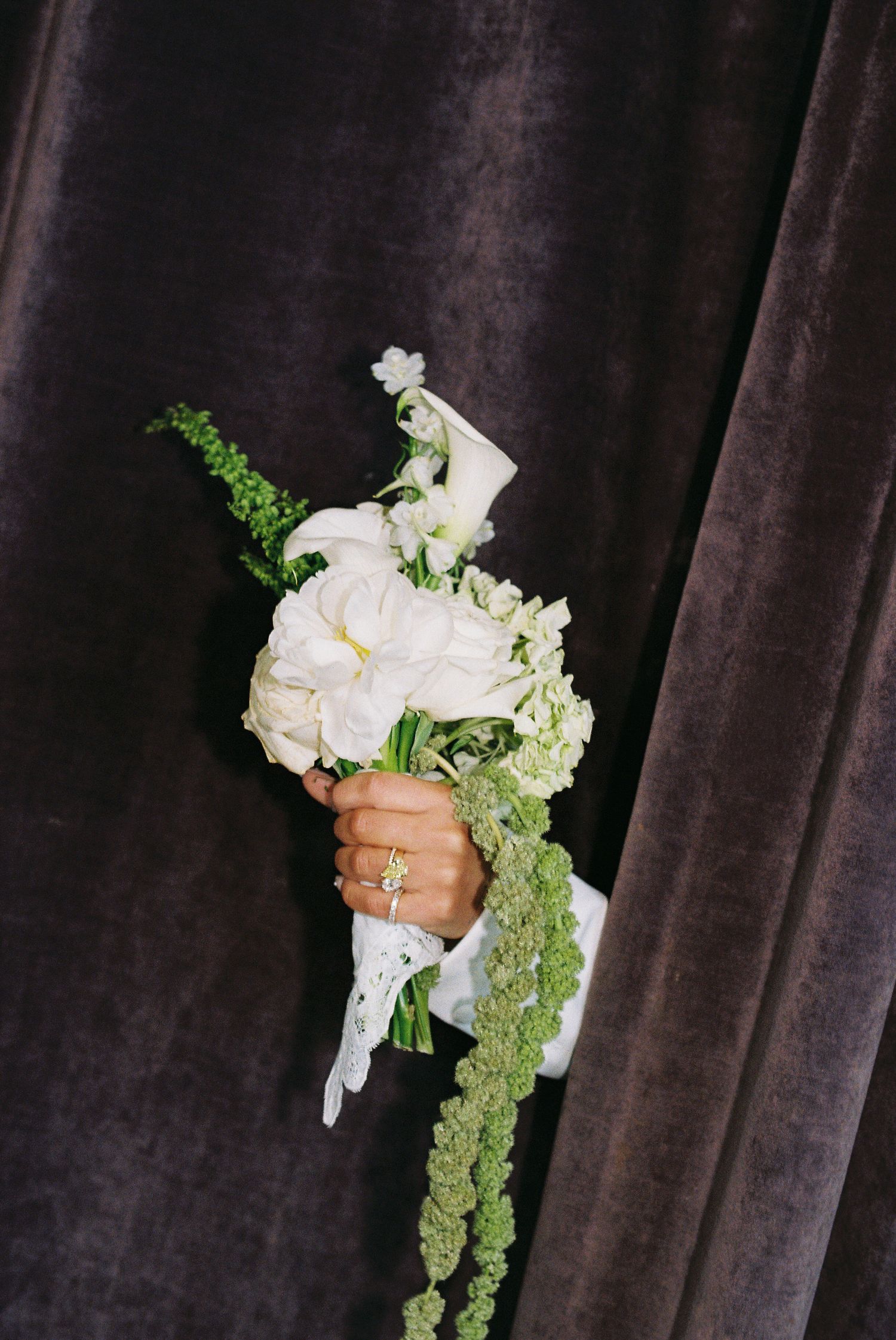 Hand holding white floral wedding bouquet with green accents against gray velvet backdrop, featuring diamond ring.