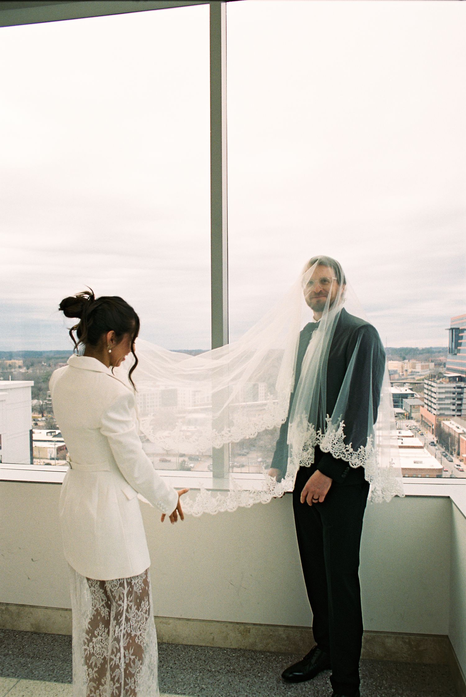 Bride and groom share a romantic first look moment, her lace veil floating between them by a high-rise window.