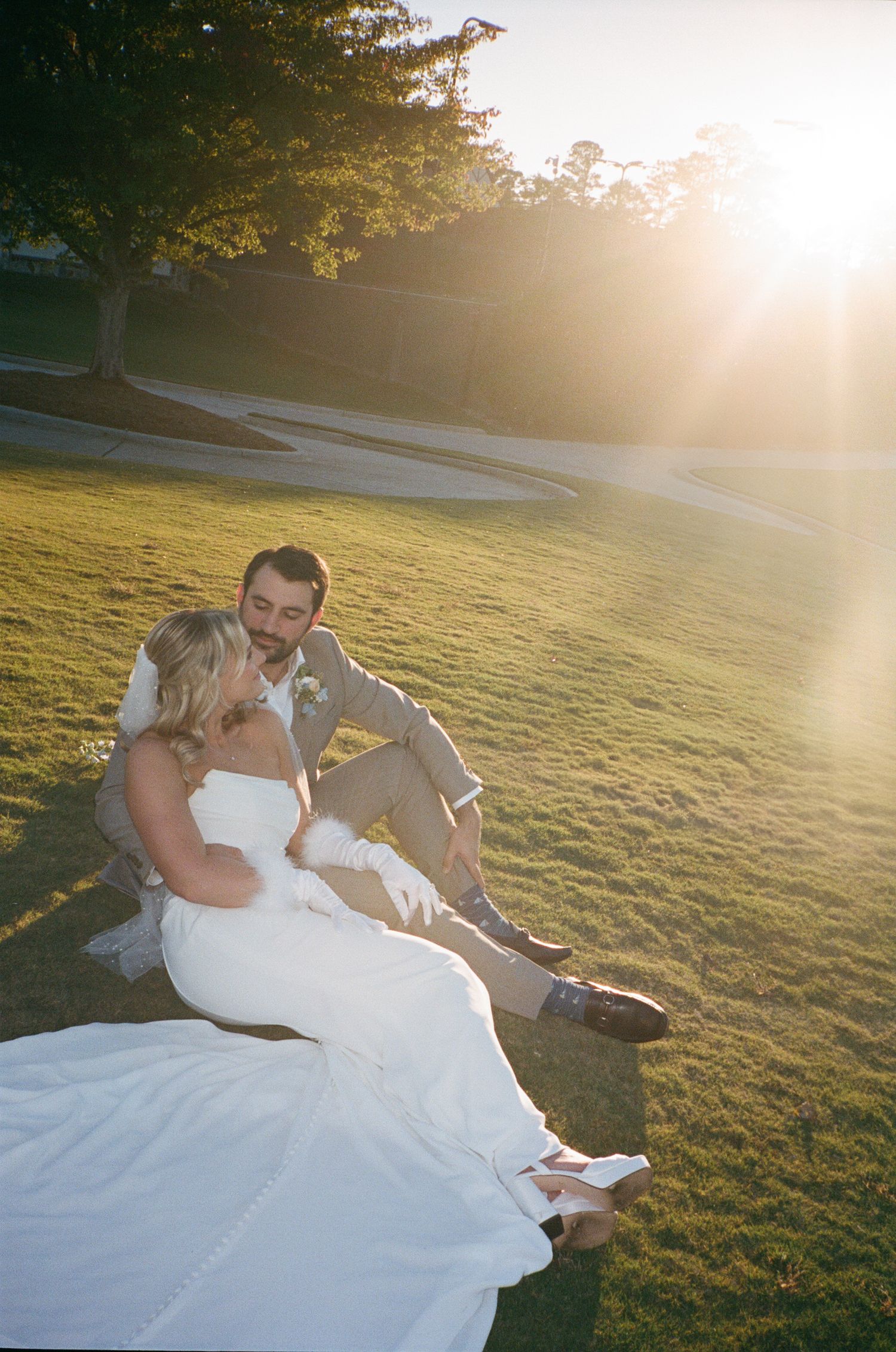 A couple in wedding attire sits together on grassy lawn during golden hour sunset.
