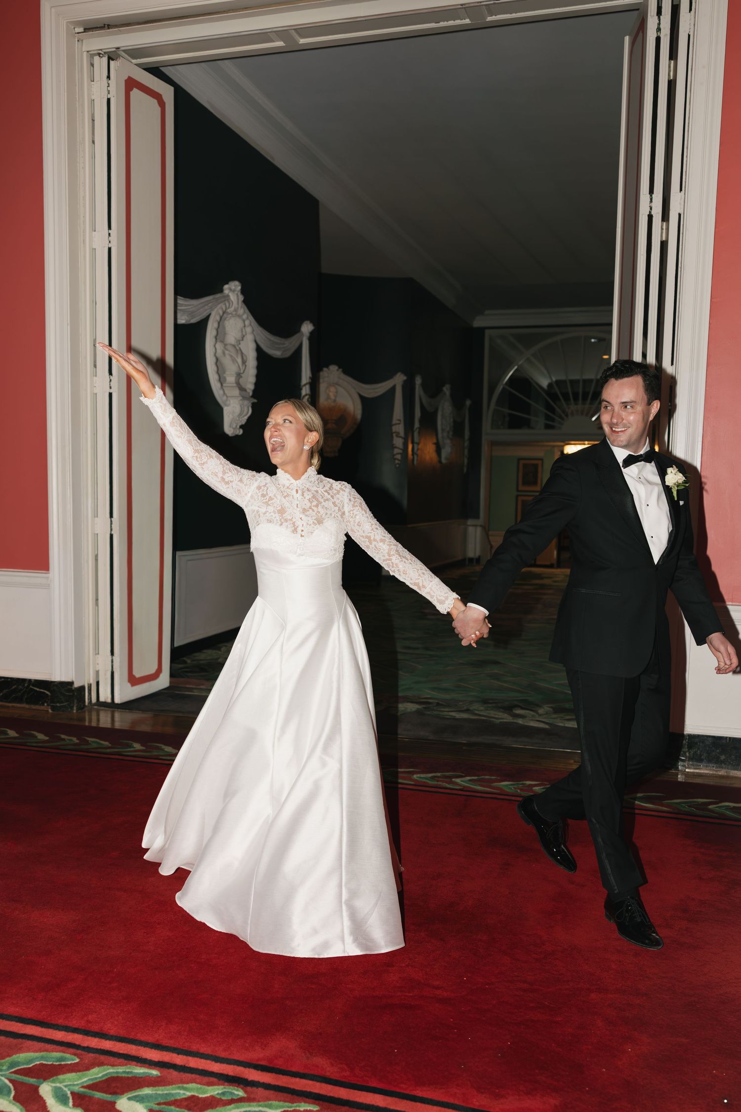 A joyful bride in white lace gown and groom in tuxedo make grand entrance on red carpet at elegant wedding venue.
