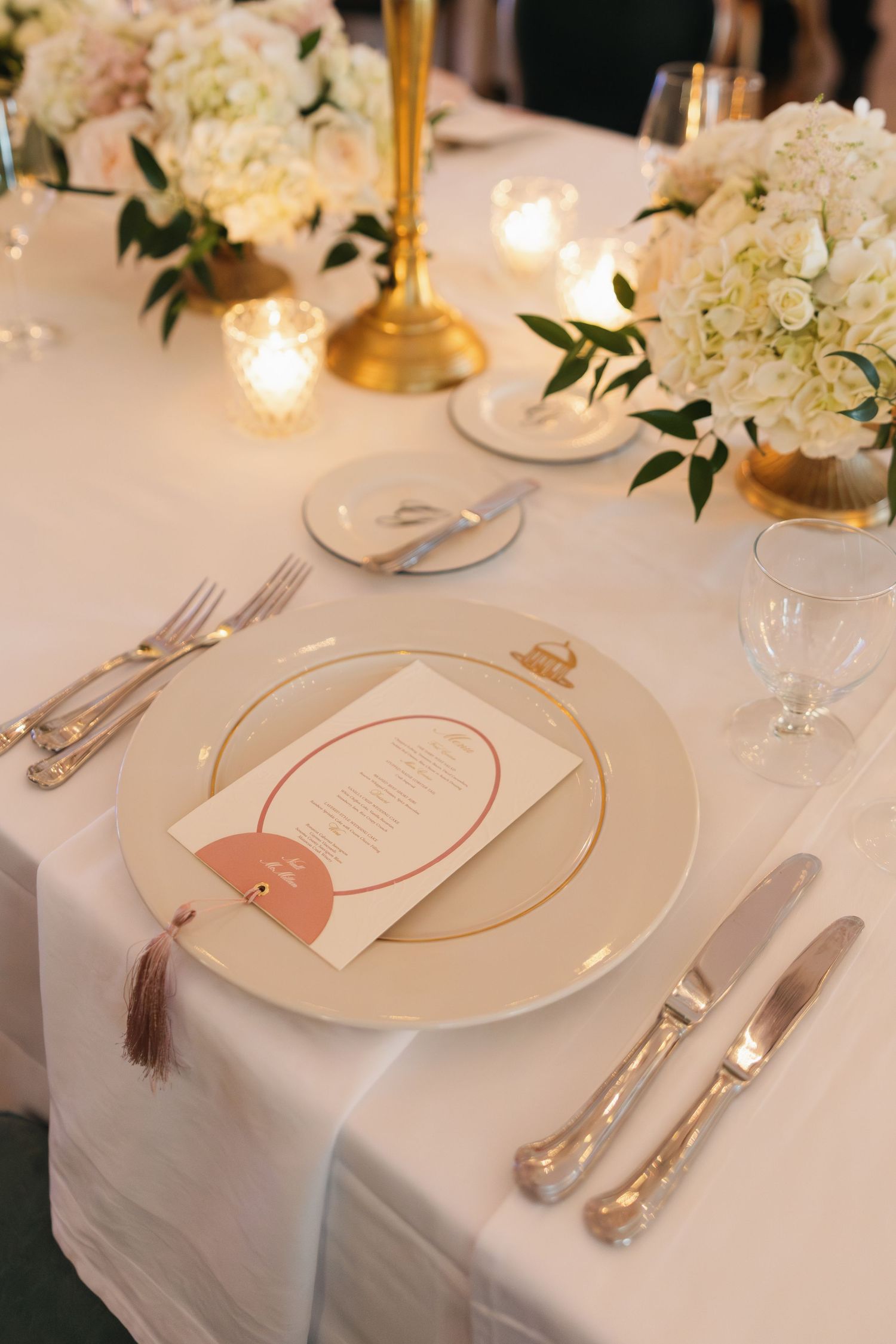 Elegant wedding table setting with white floral centerpieces, gold charger plates, silver cutlery, and a blush menu card.