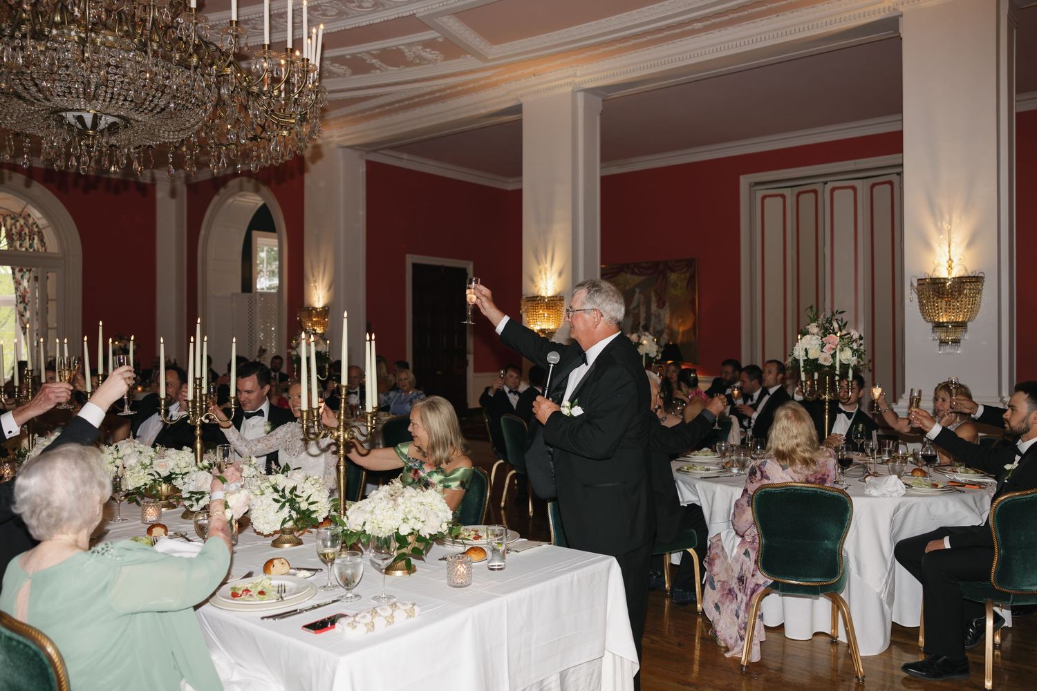 Elegant formal dinner banquet with guests raising glasses for a toast in a grand ballroom with chandeliers.